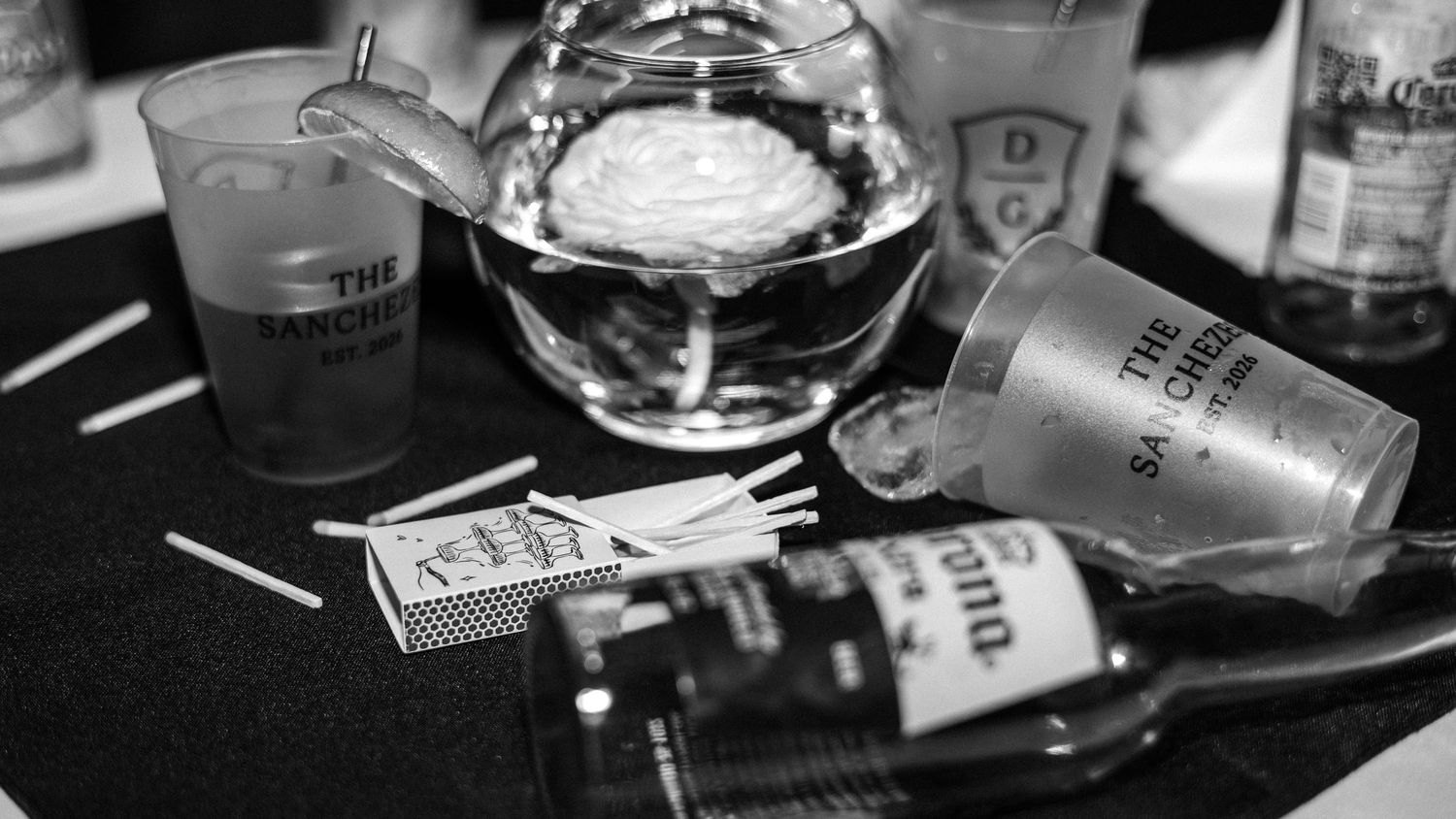 Black and white photo of overturned cups, matchbook, and liquor bottles on a dark table.