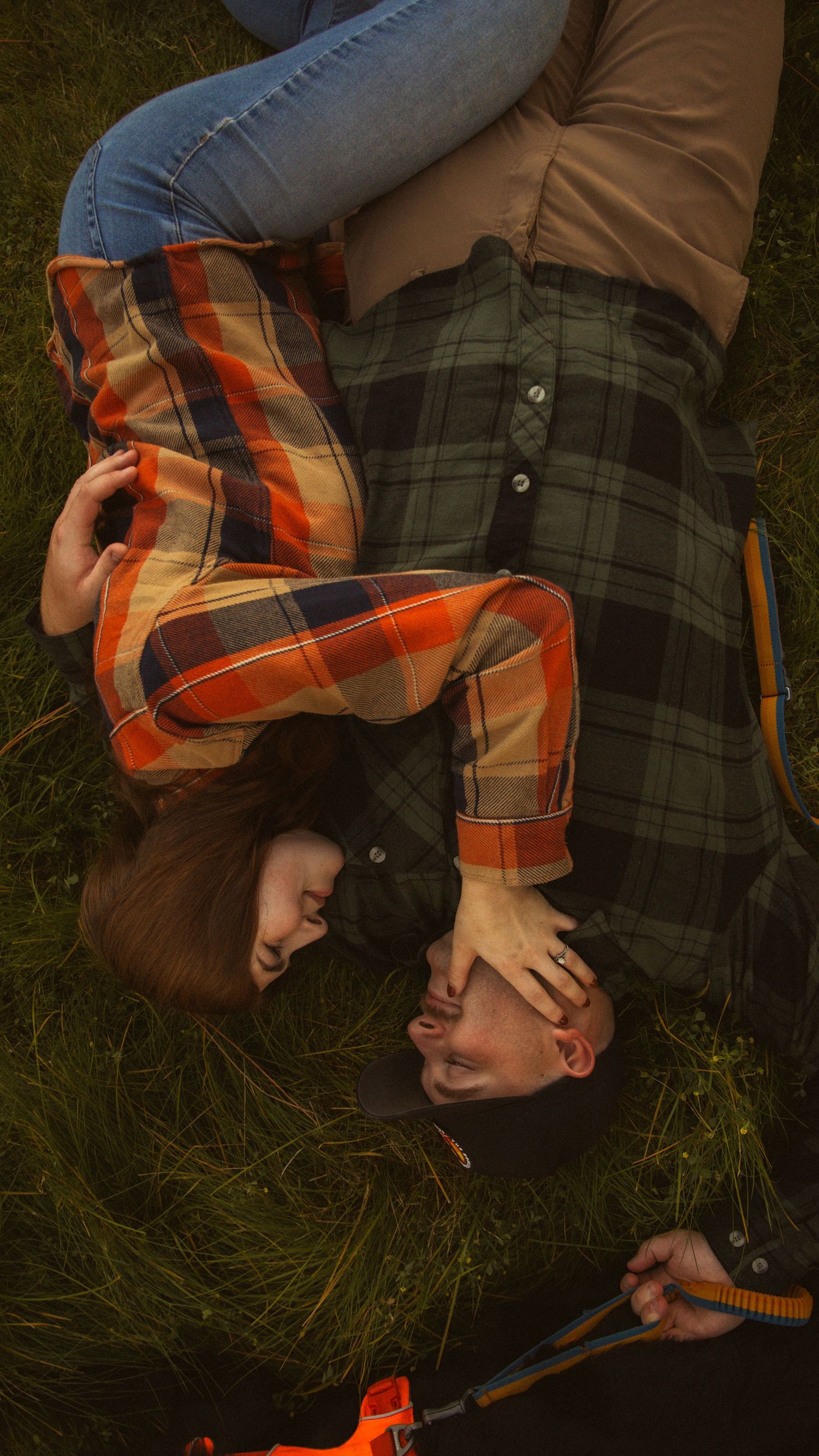 Couple lying on grass in plaid flannel shirts, smiling and embracing in a romantic outdoor autumn setting.