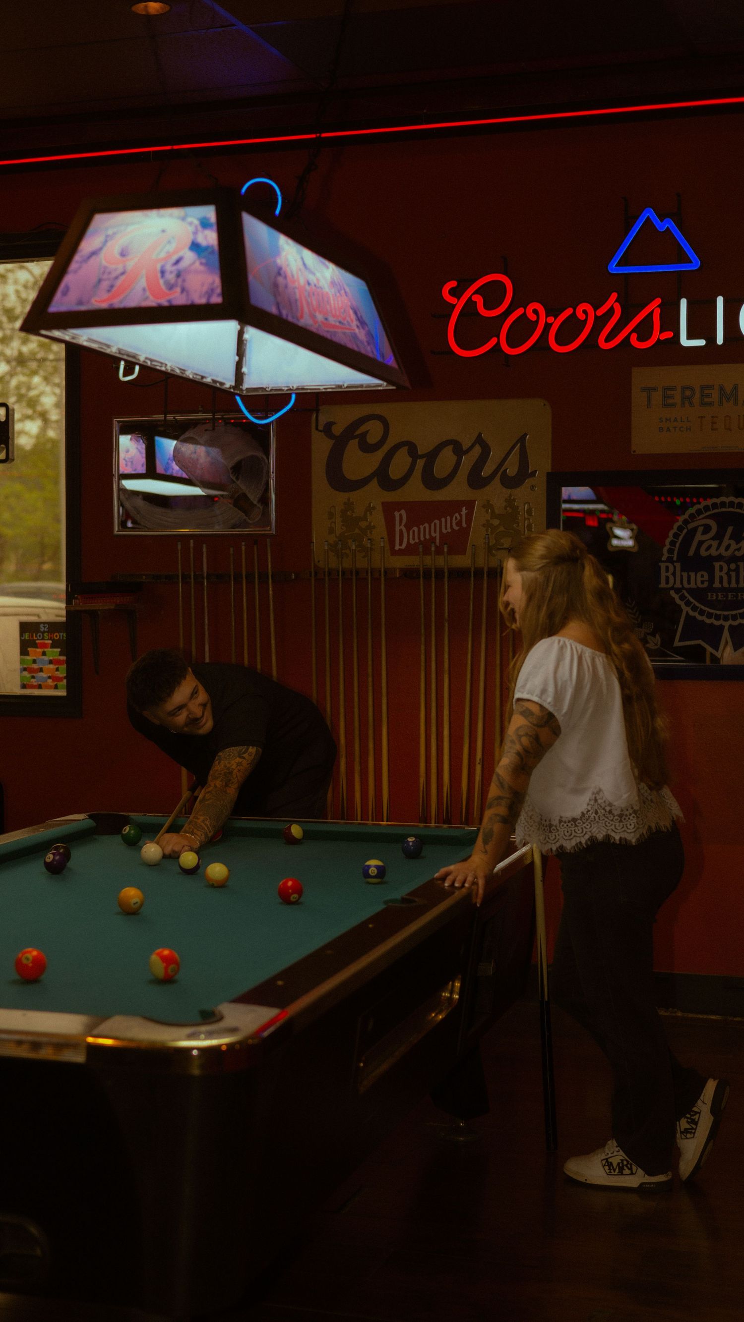 Two people playing pool in a dimly lit bar with Coors Light neon signs and vintage beer advertisements on red walls.