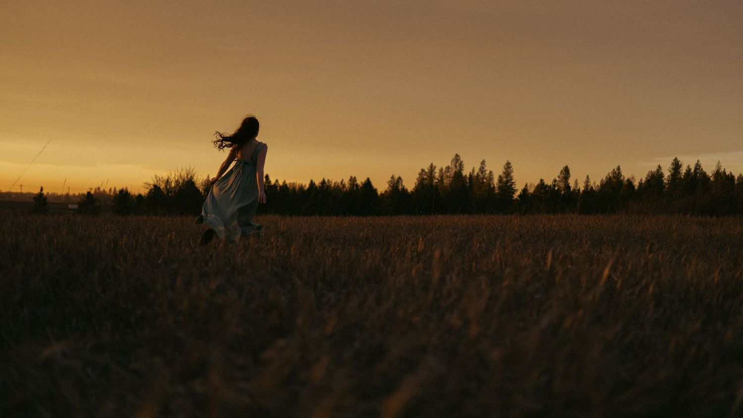 A woman in a flowing dress runs through a golden field at sunset with a forest silhouette in the background.