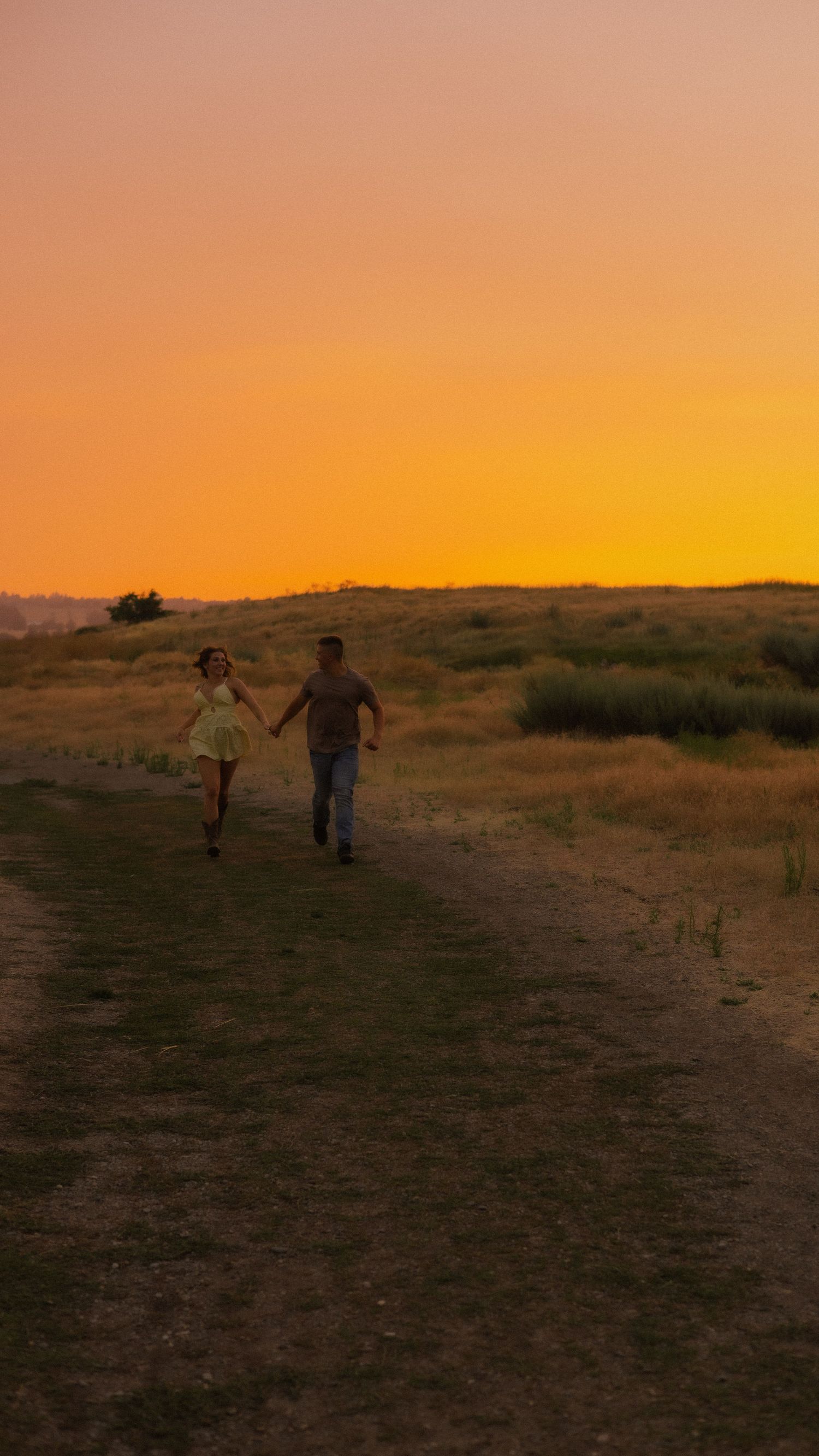 Couple holding hands running on a dirt path through golden fields at sunset with vibrant orange sky.