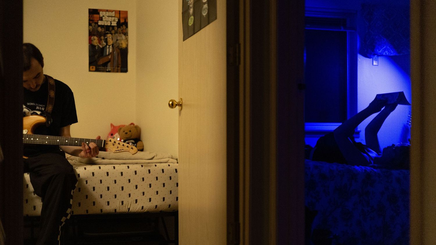 A person plays guitar in a dimly lit room while blue light illuminates an adjacent bedroom through a doorway.
