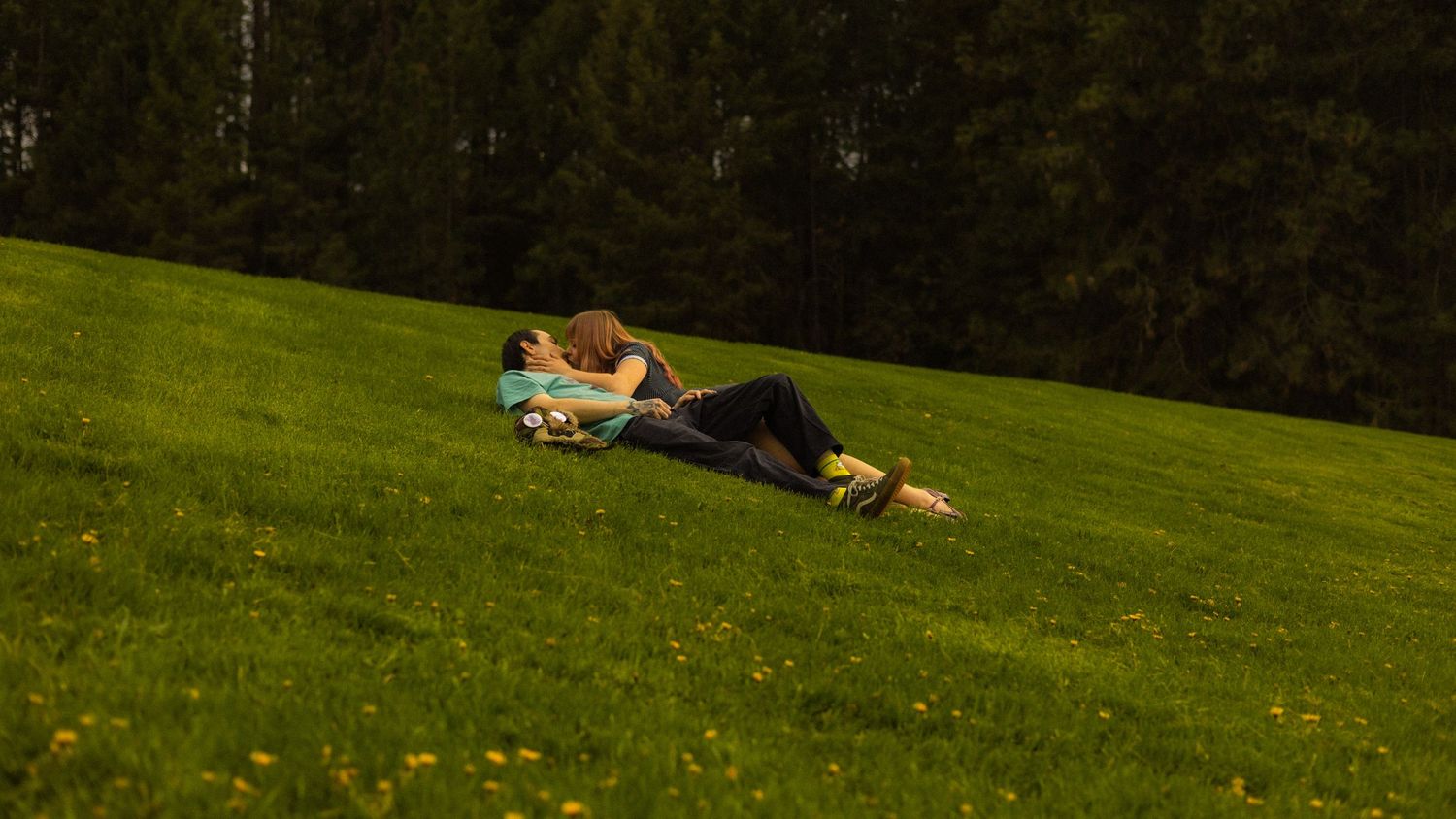 A couple lying together on a sunny green hillside meadow surrounded by wildflowers.
