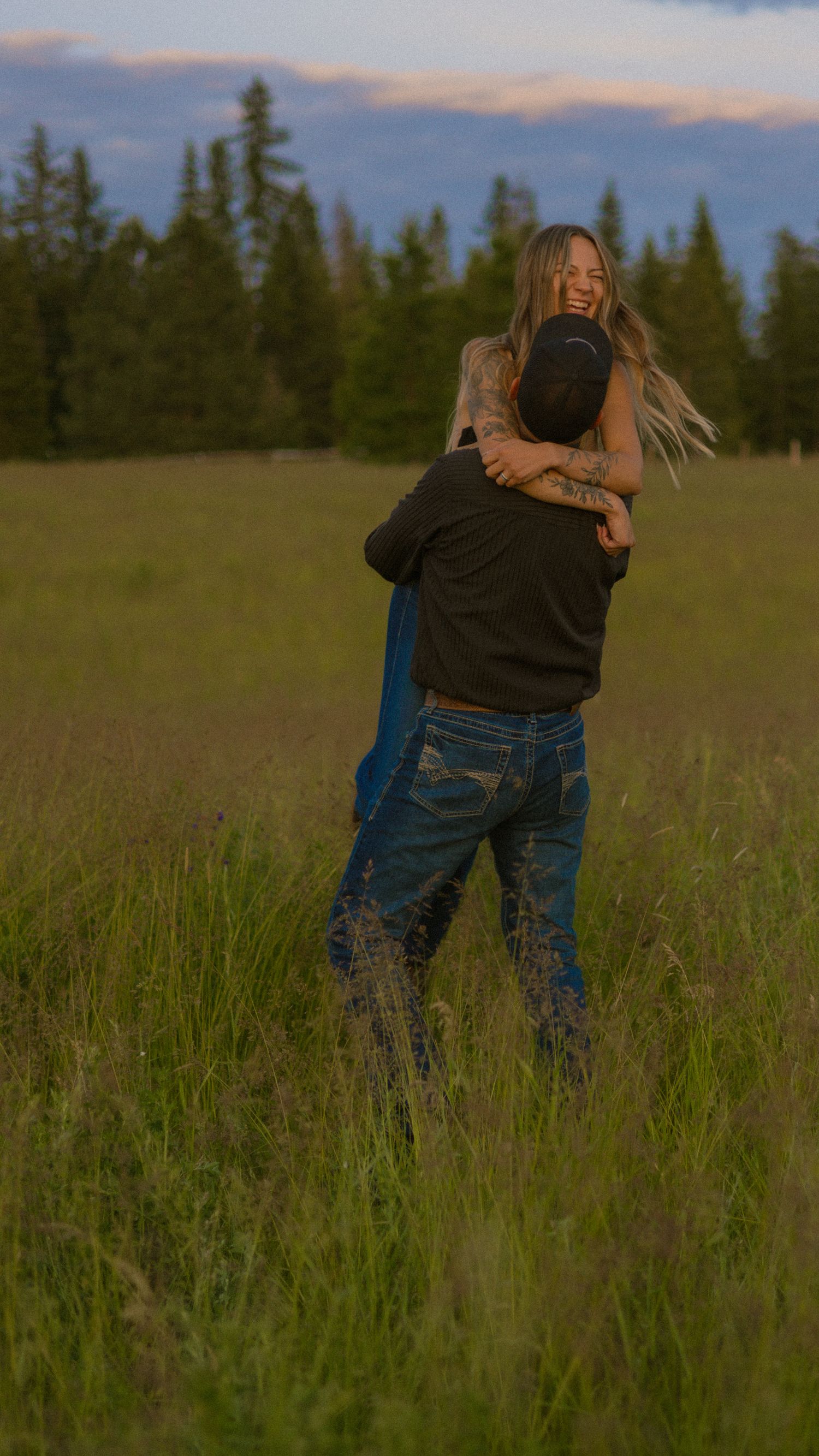 Couple embracing and laughing in a green meadow at dusk with pine trees and colorful sky in background.