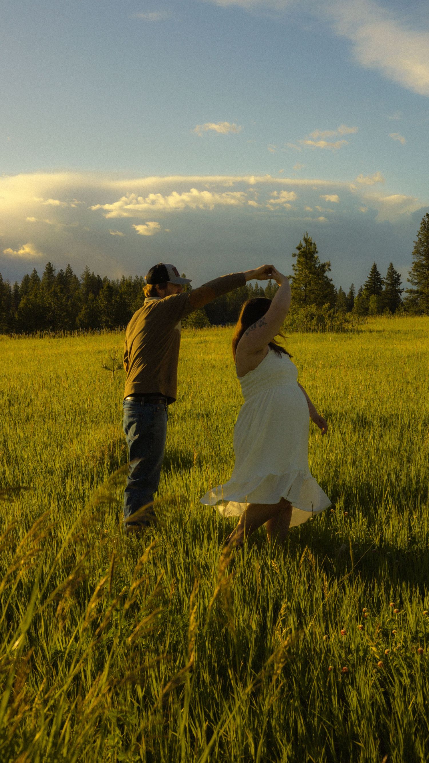Couple dancing in a lush green meadow at golden hour, woman in white dress being twirled by man in jeans.