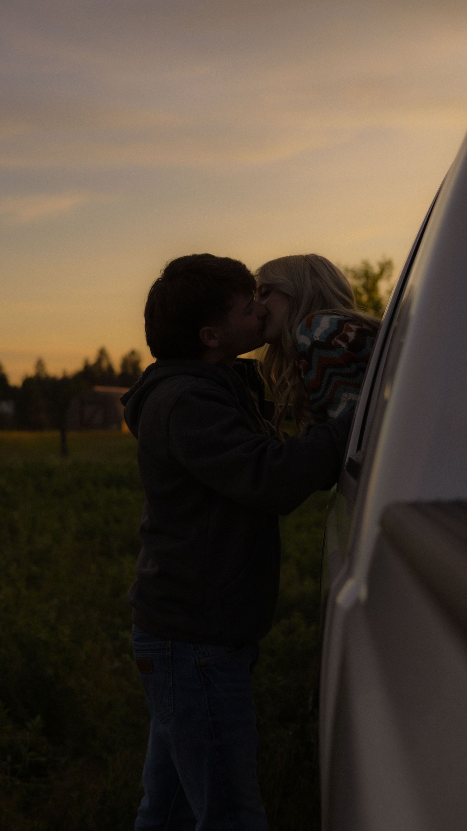 A couple shares a tender kiss beside a white vehicle in an open field during a golden sunset.