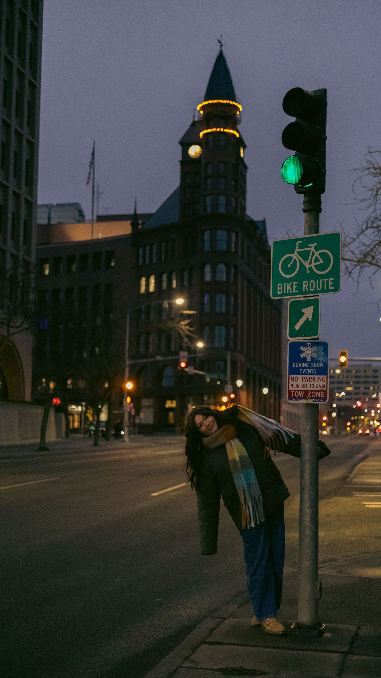 A woman leans against a bike route sign post on an empty city street at dusk near a historic tower.