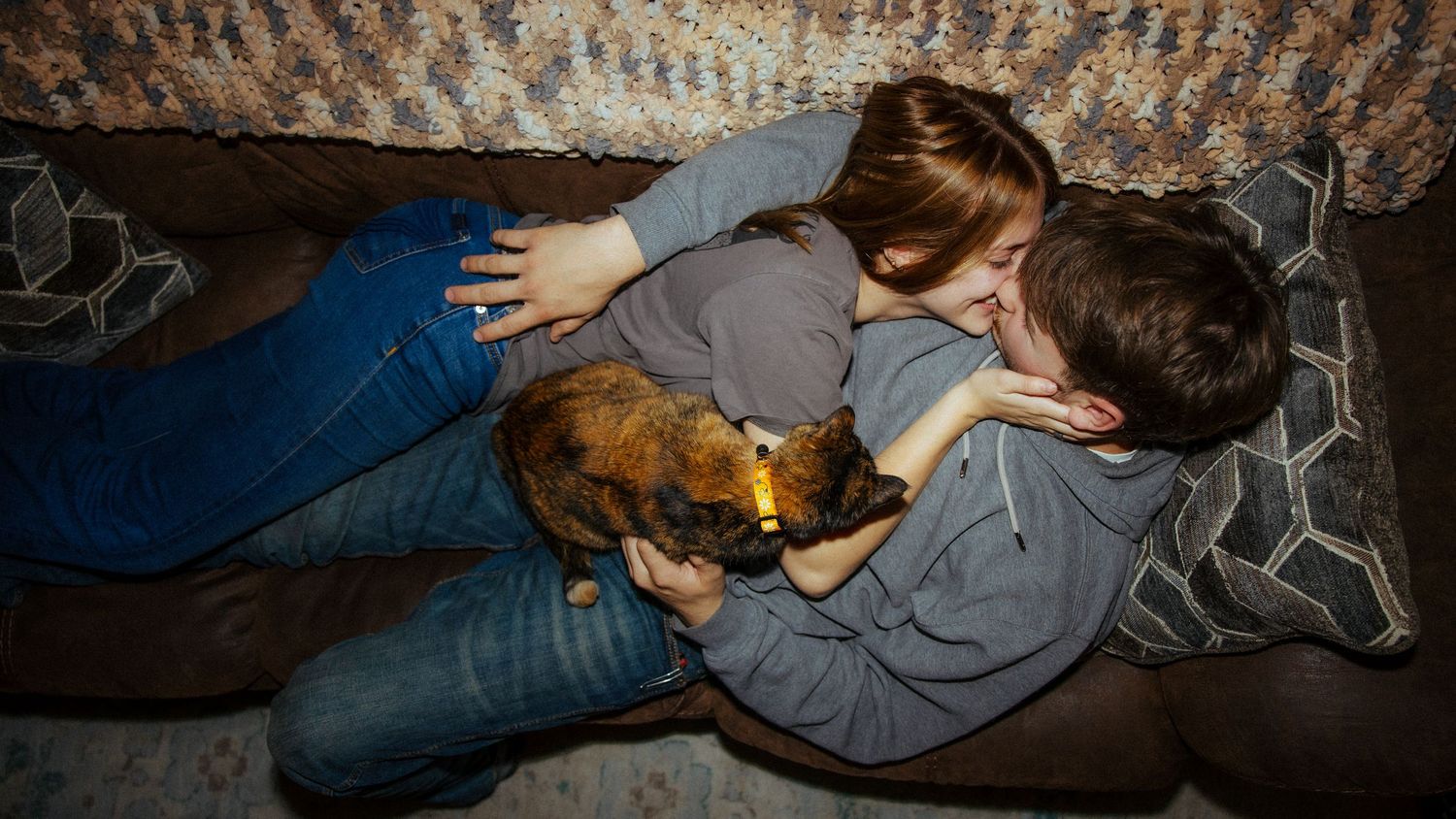 Two children cuddle with a small cat on a couch in a dimly lit room.
