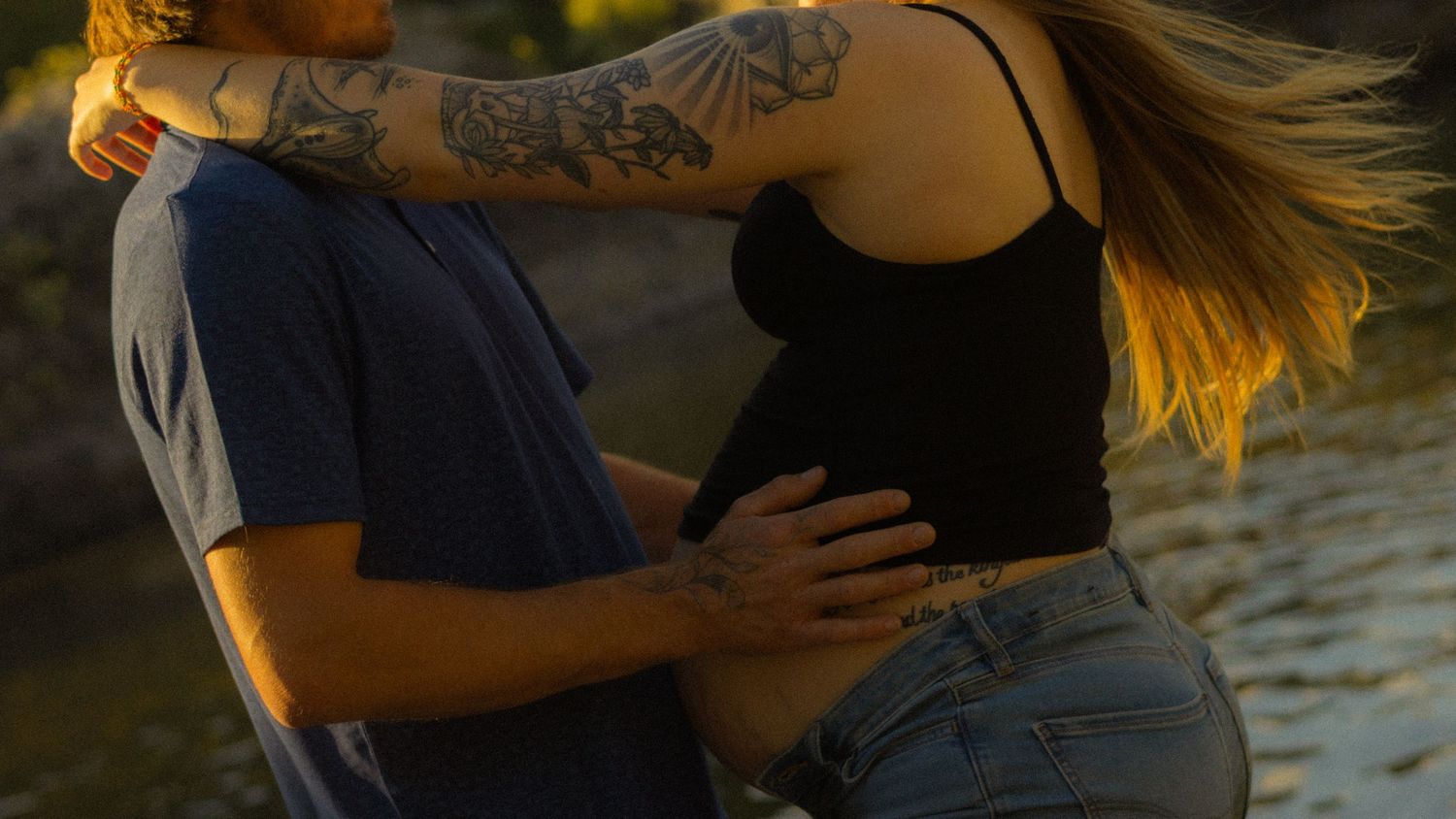 Couple embracing outdoors at golden hour, tattooed man touching pregnant woman's belly near water.