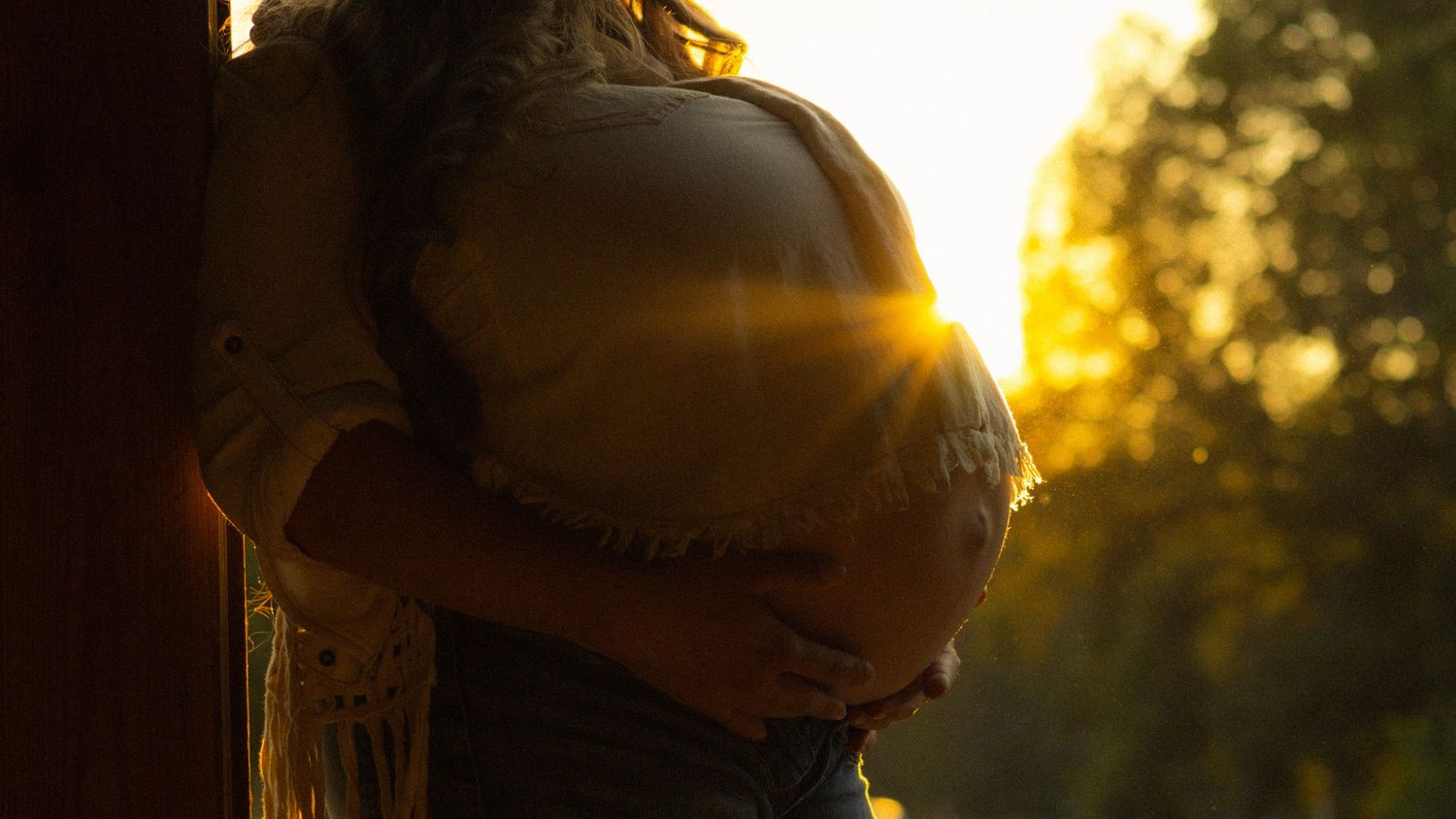 A pregnant woman cradles her baby bump outdoors in golden sunset light filtering through trees.