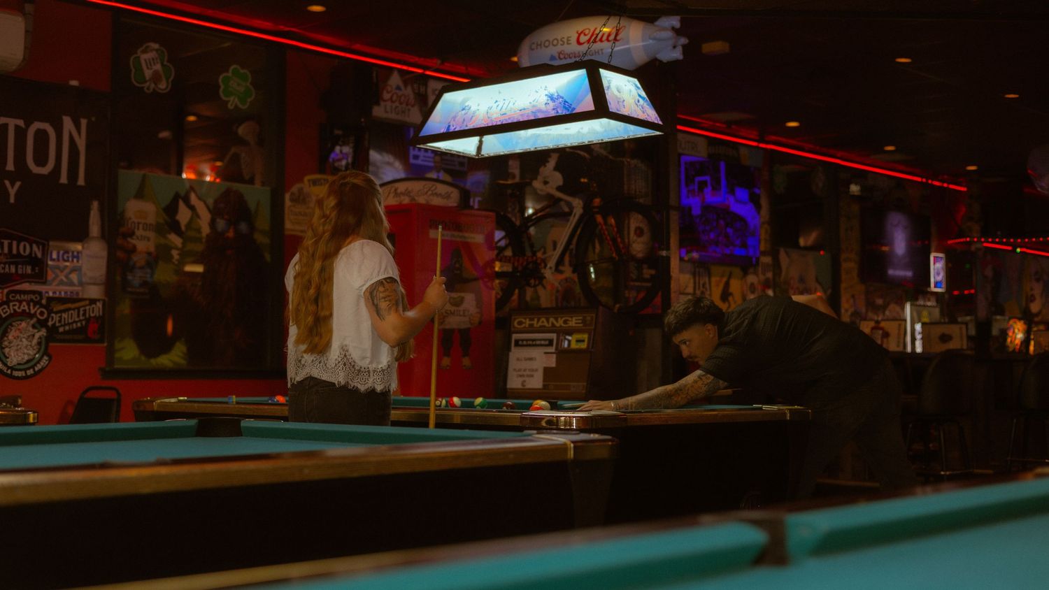 Two women playing pool in a dimly lit bar with neon signs and a classic billiards table lamp overhead.
