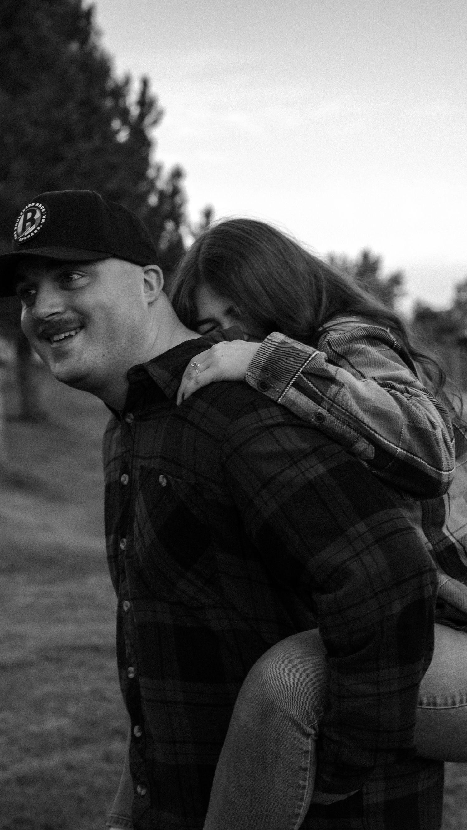 Black and white photo of a smiling man in a plaid shirt and cap giving a young girl a piggyback ride outdoors.
