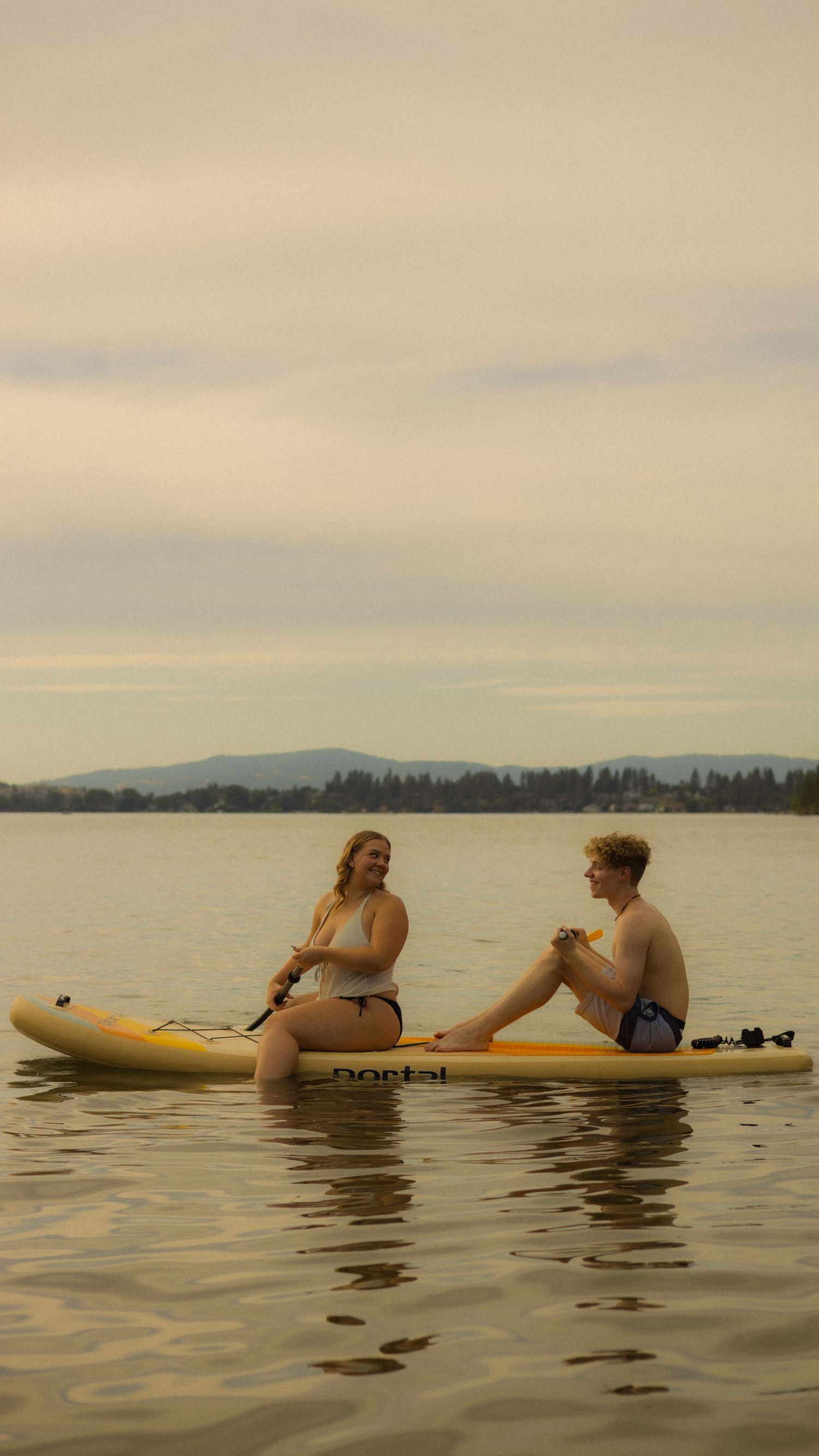 Two people sit on a yellow paddle board on calm water with mountains and trees in the background.