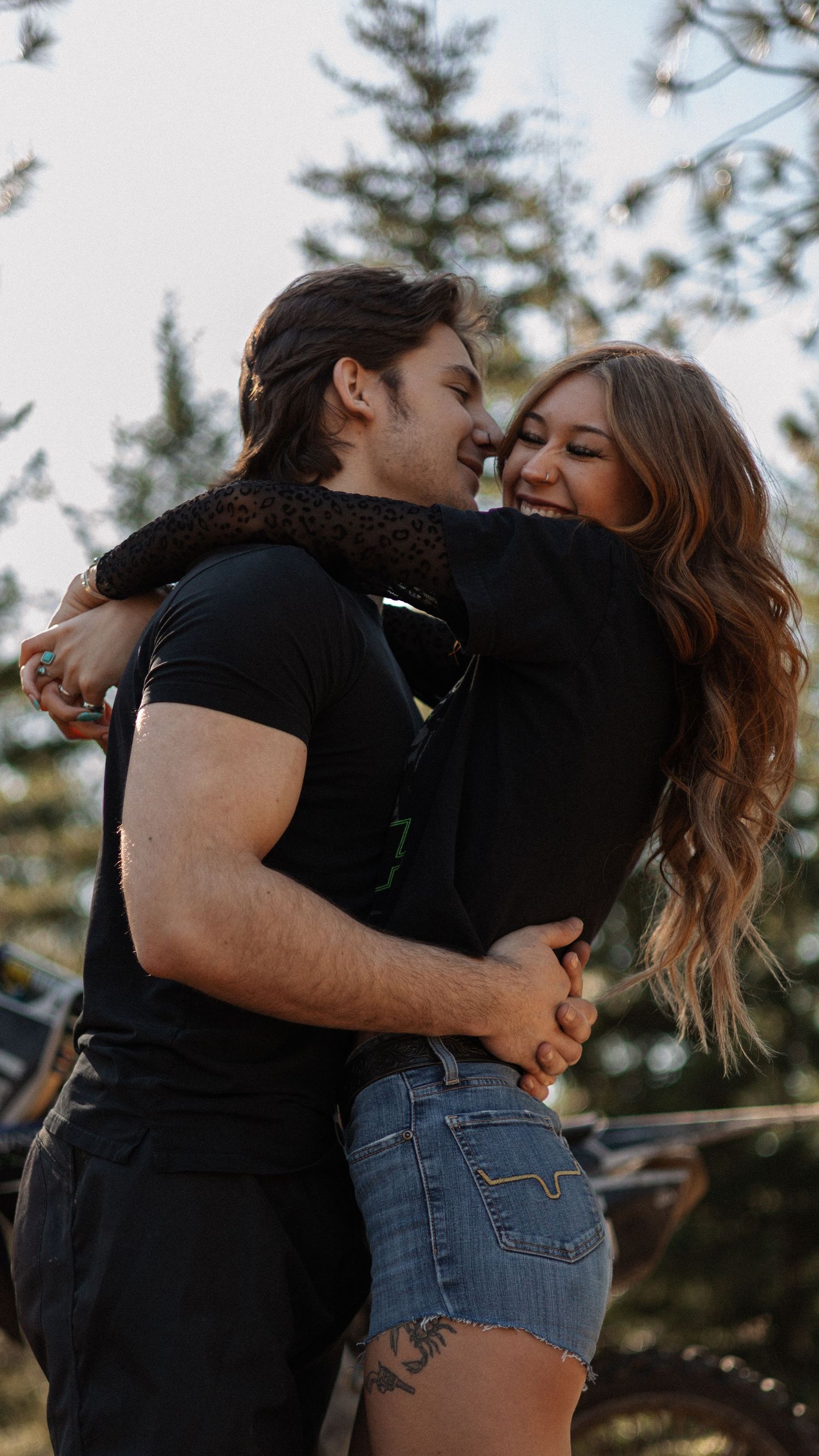 A smiling couple embraces joyfully outdoors near pine trees, both wearing black shirts, the woman in denim shorts.