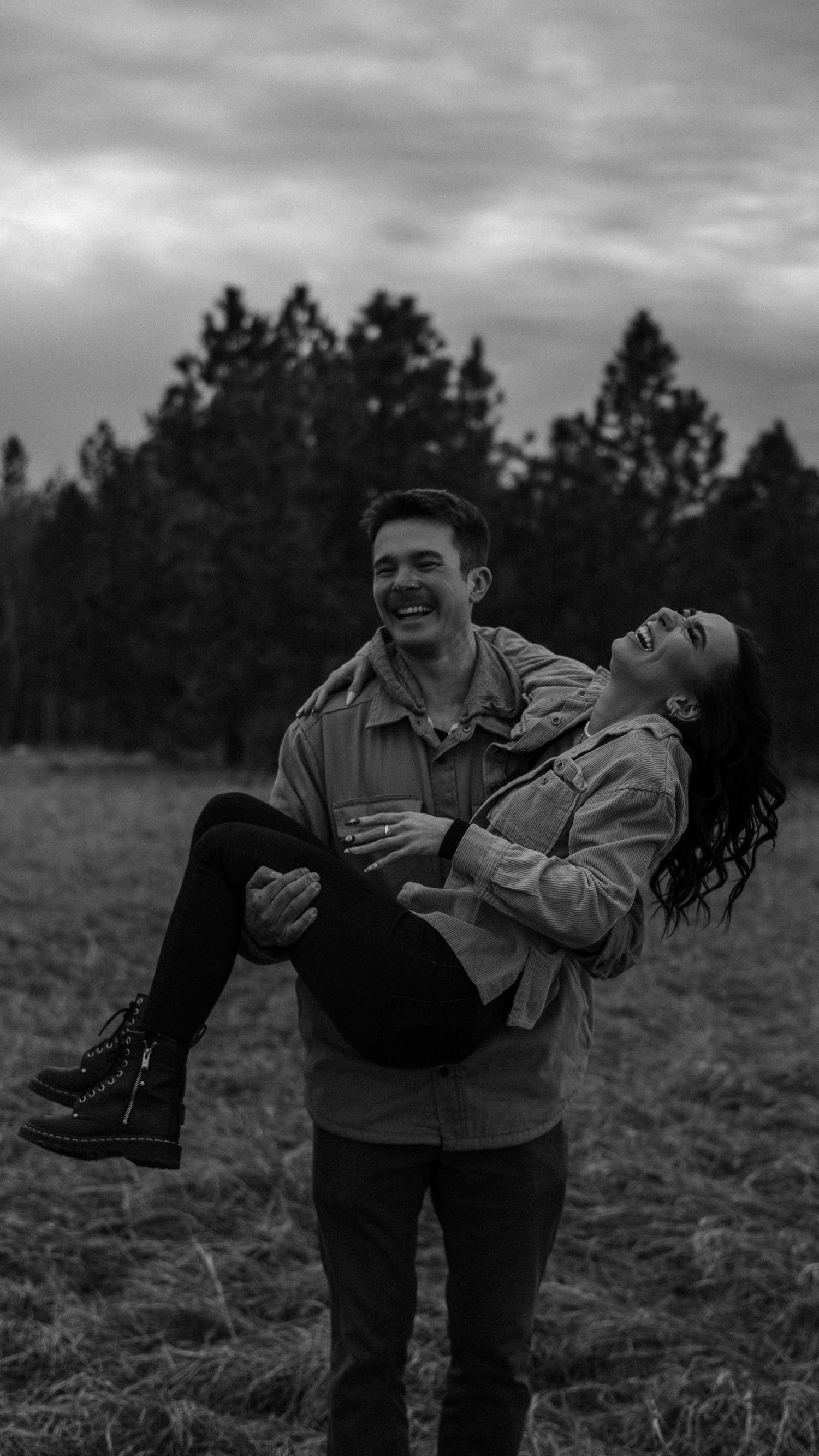 Black and white photo of a man carrying a laughing woman in his arms in a field with trees behind them.