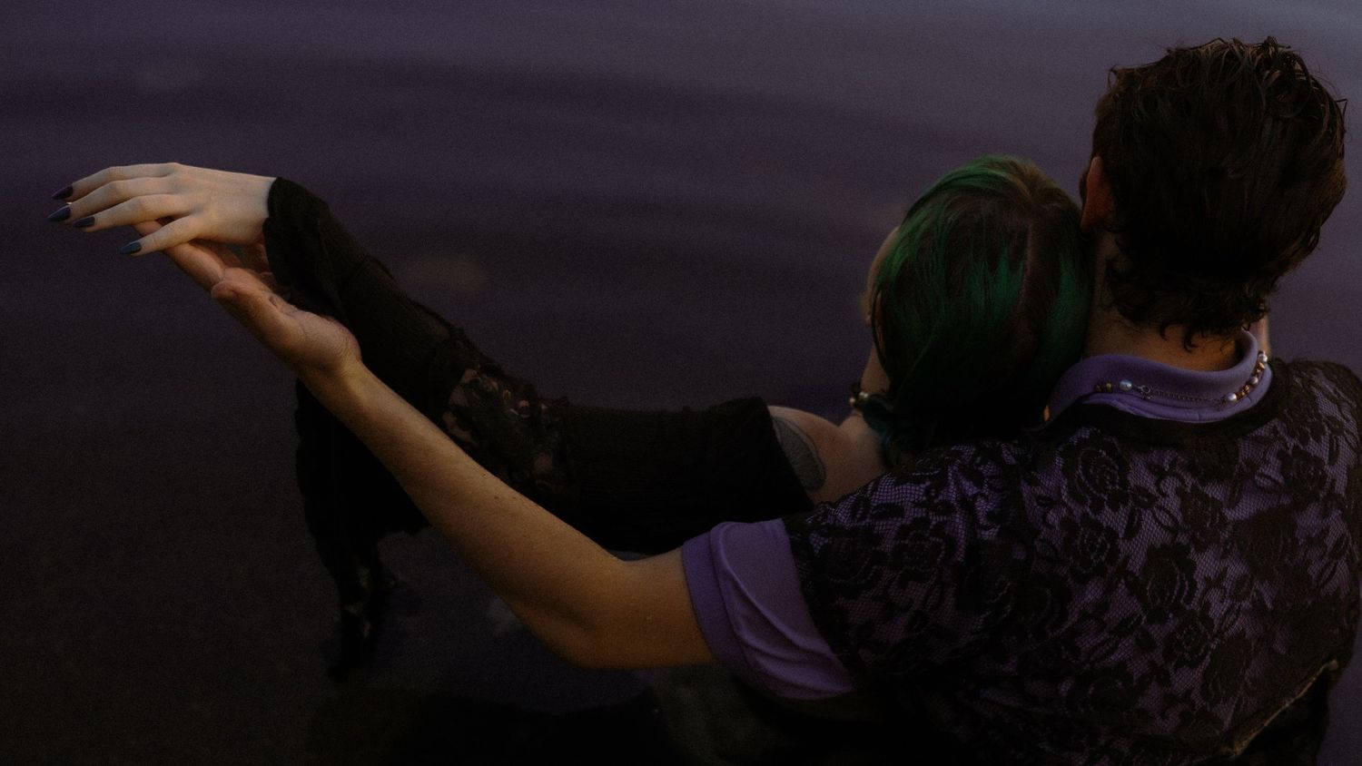 Couple sitting together by dark water, one with green hair, embracing romantically at dusk.