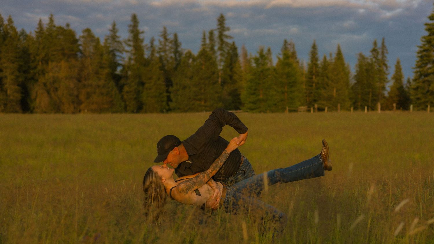 Couple embracing and playing in a lush green meadow with tall evergreen trees under a dramatic cloudy sky.