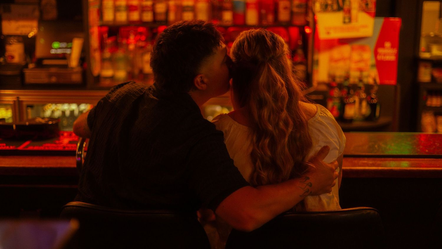Couple sharing a romantic kiss at a dimly lit bar with warm red and orange lighting.