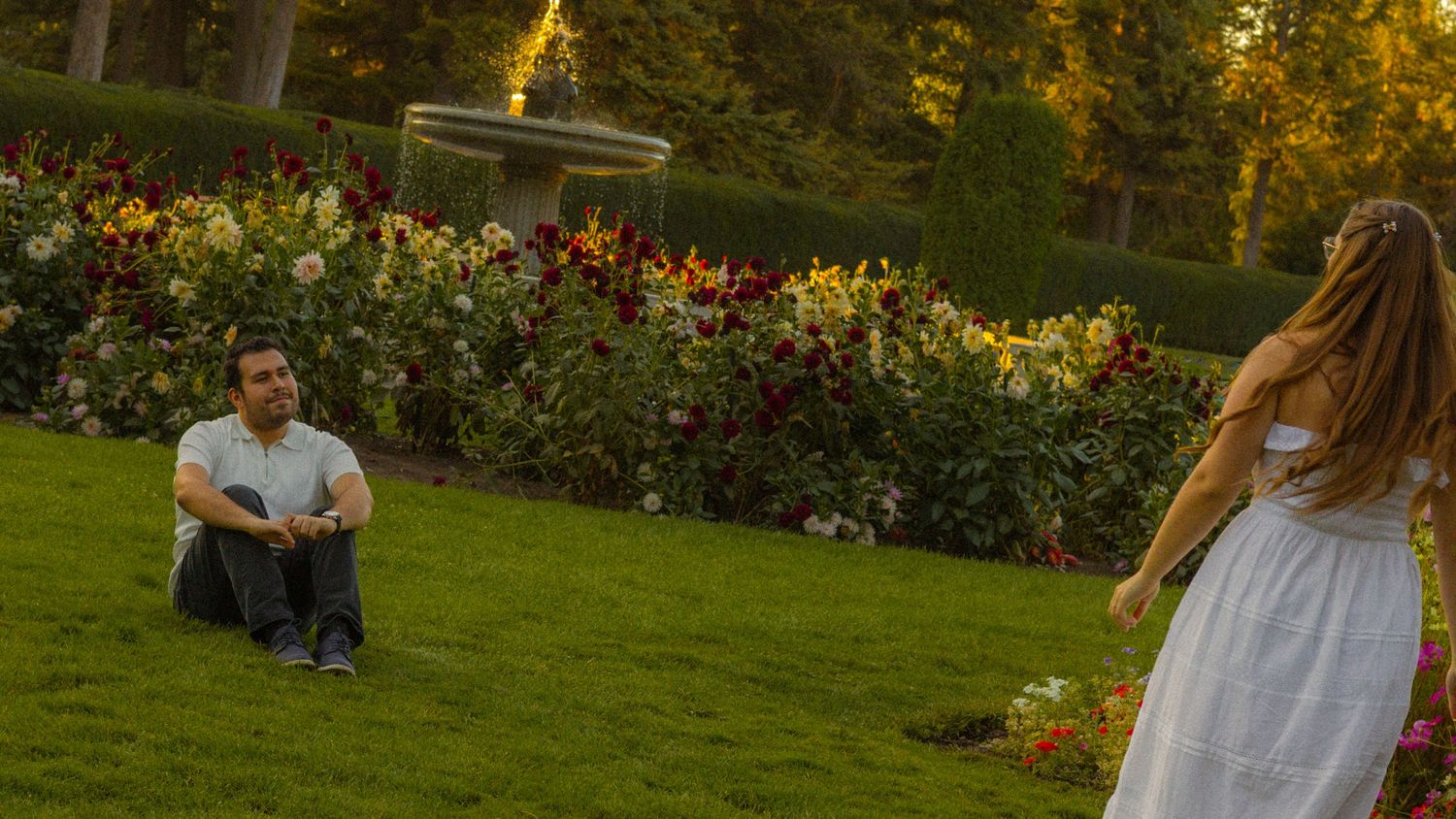 Man sitting on grass in a garden watching a woman in a white dress near colorful flower beds.