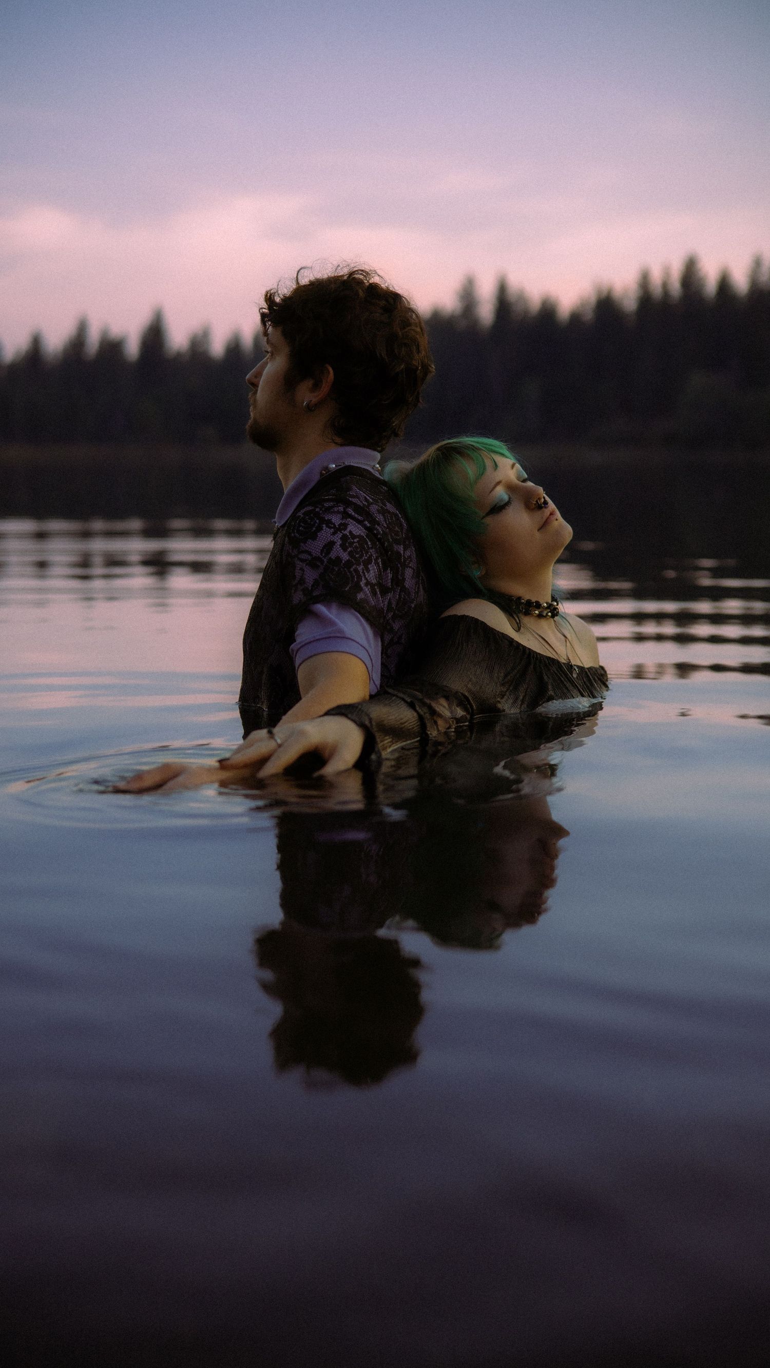 Two people in dark clothing stand waist-deep in a calm lake at dusk, with pine trees and purple sky reflected on water.