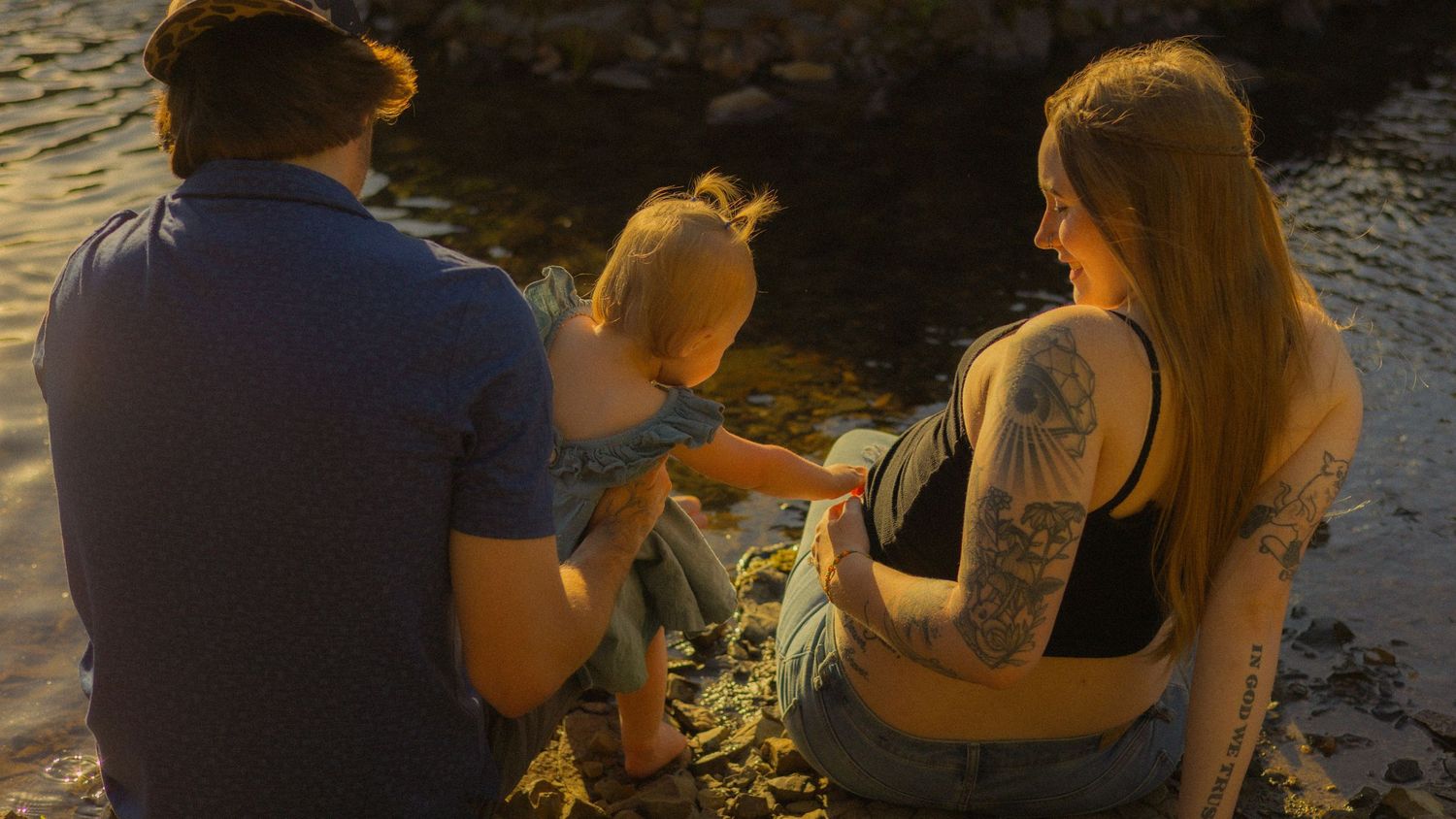 A tattooed family with a baby sits by a sunlit rocky shoreline during golden hour.