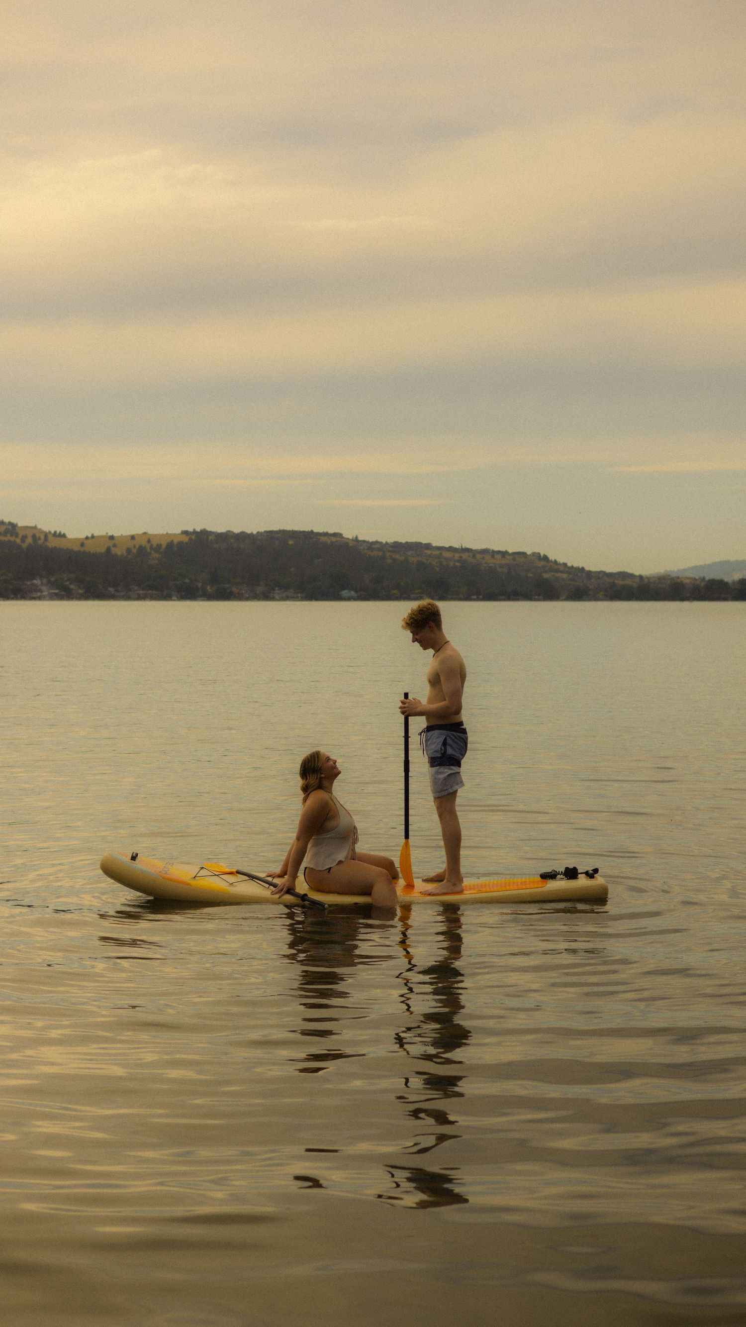 Two people on a yellow inflatable paddleboard at sunset on a calm lake with hills in the background.