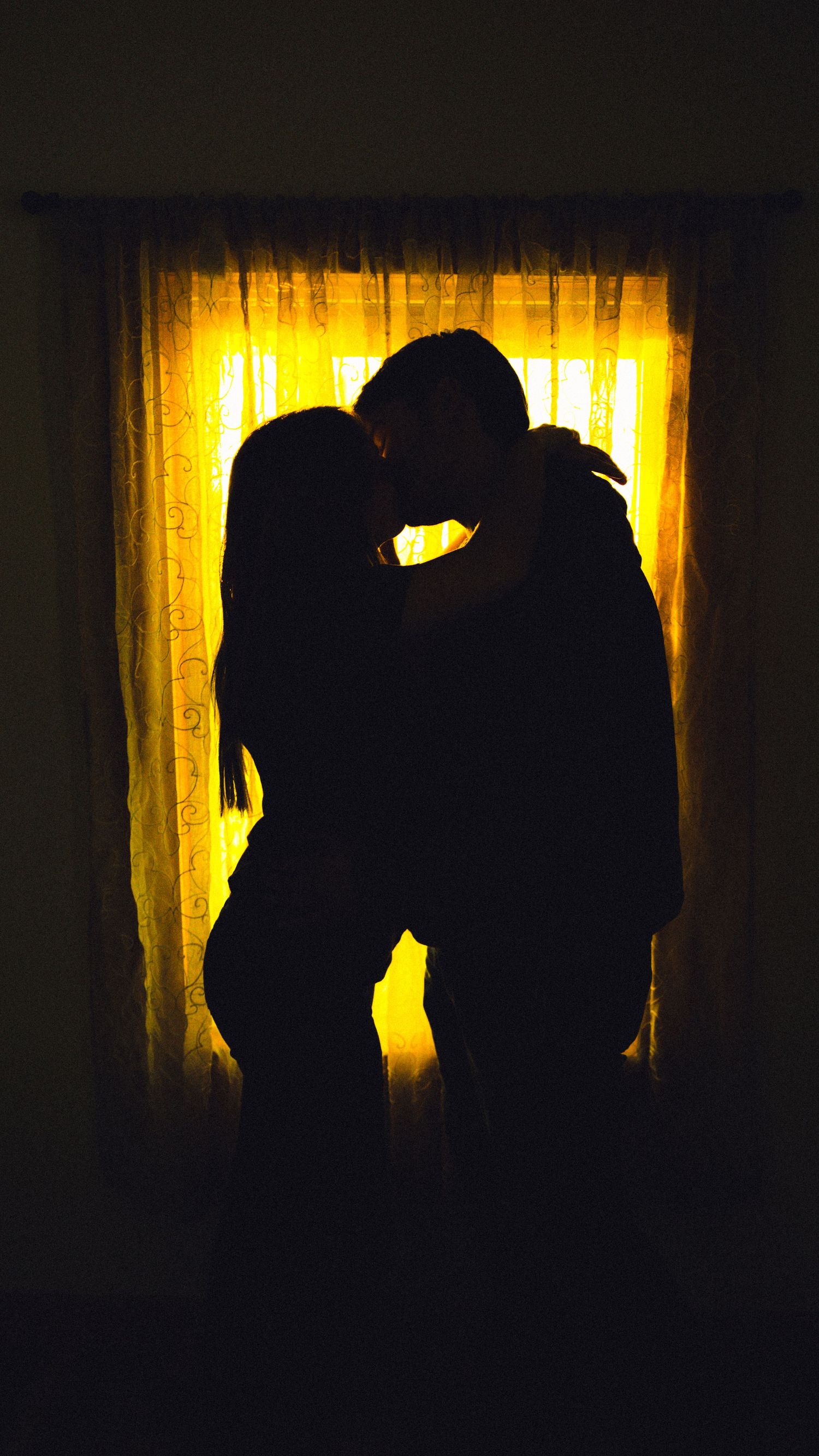 Romantic silhouette of a couple kissing in front of a glowing golden window with sheer curtains in a dark room.