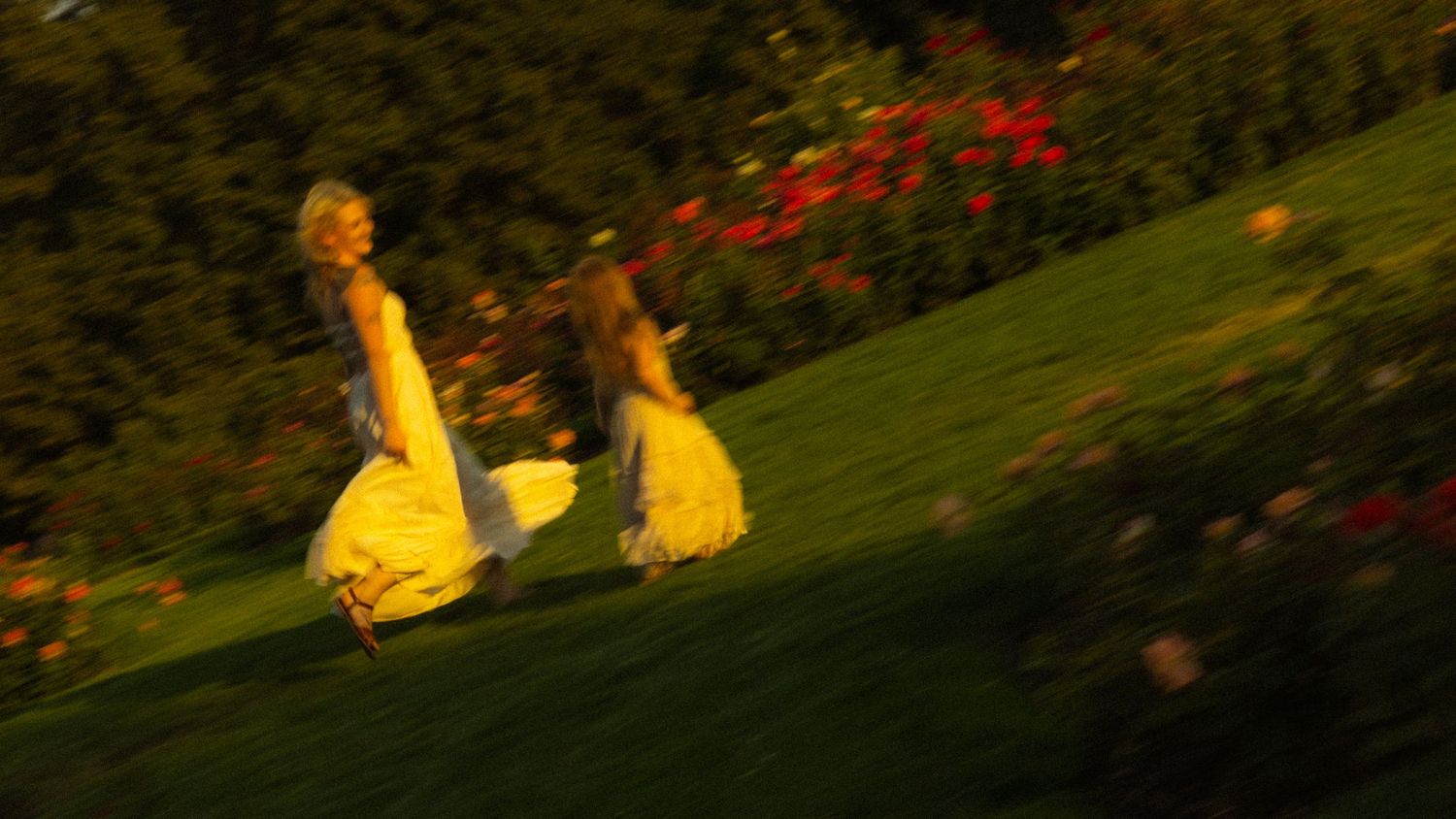 Two women in yellow dresses running joyfully across a lush green hillside garden with colorful flowers.