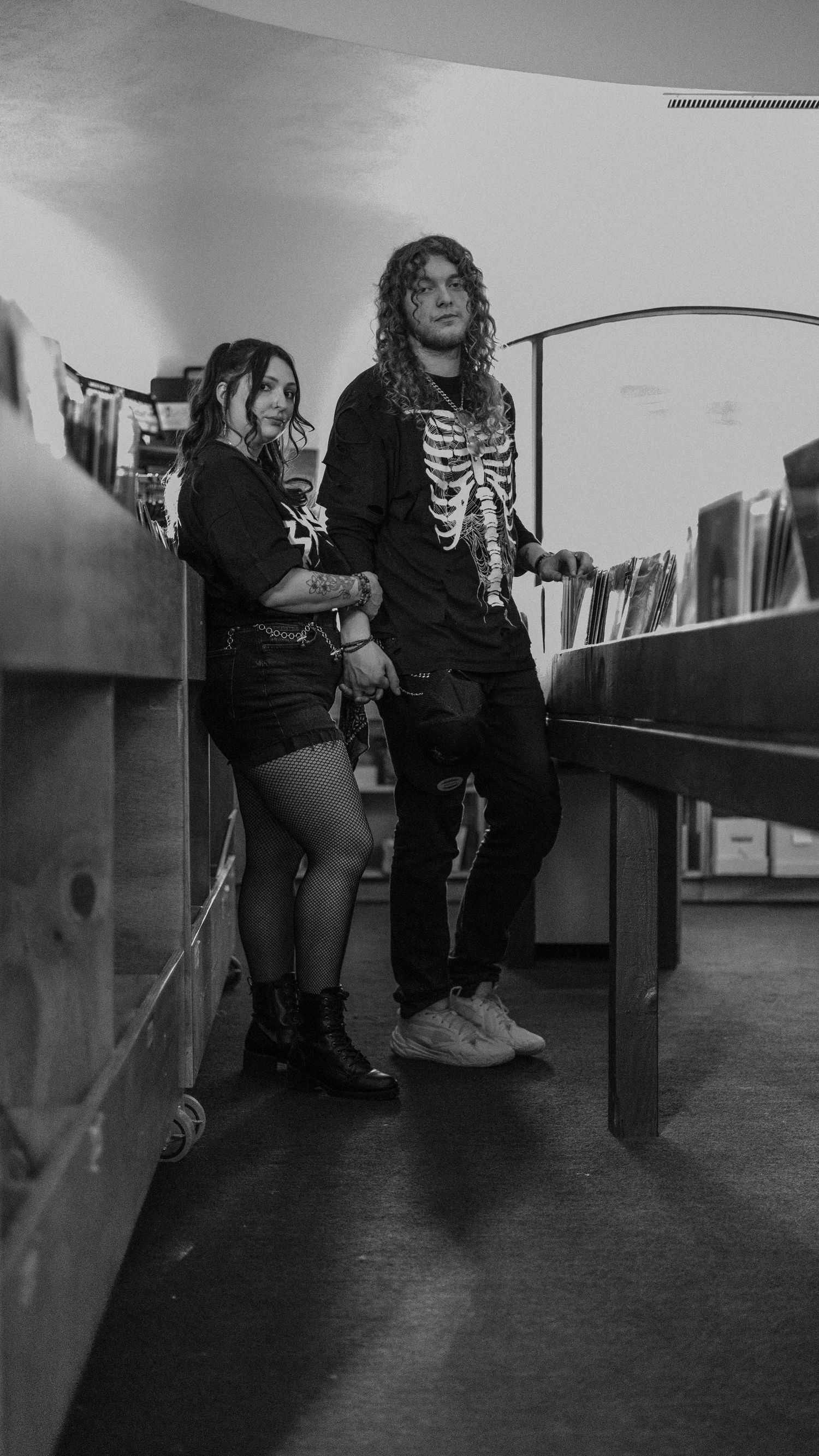 Black and white photo of two alternative style young adults standing between record bins in a vinyl music store.