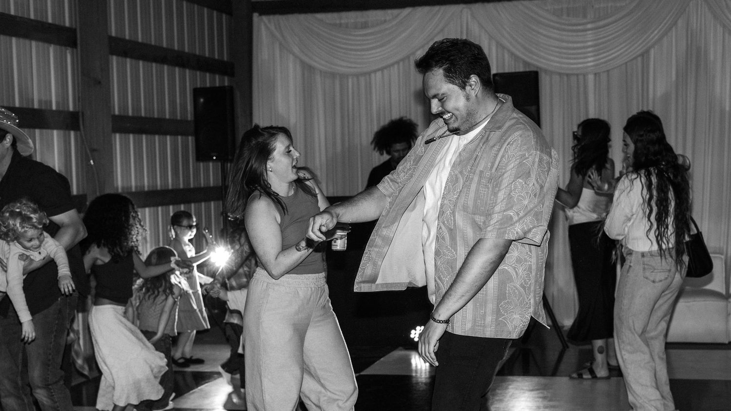 Couple dancing together at a lively party in black and white photo with guests celebrating in background.