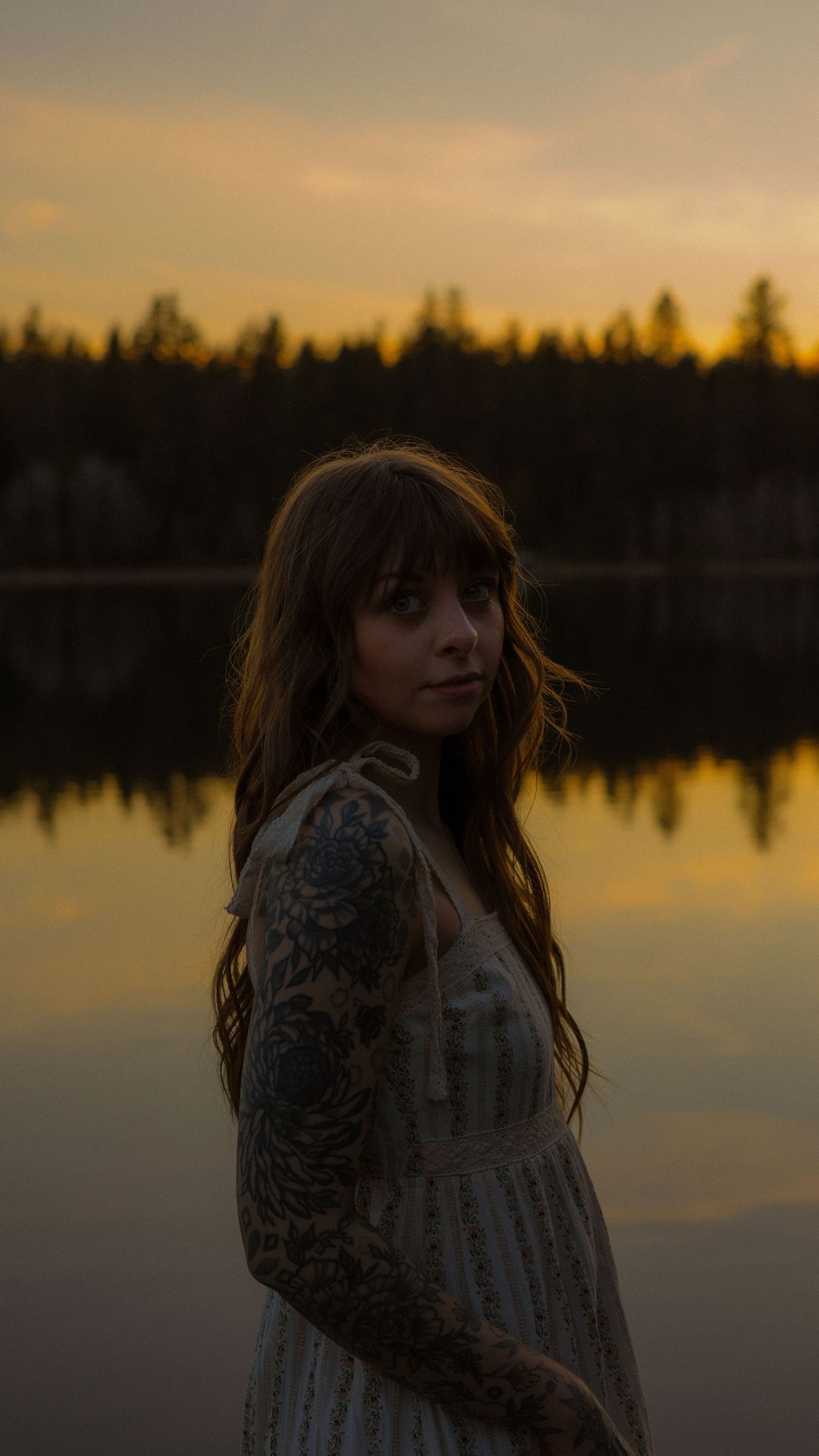 A tattooed woman in a white dress stands by a reflective lake at golden hour with forested hills behind her.