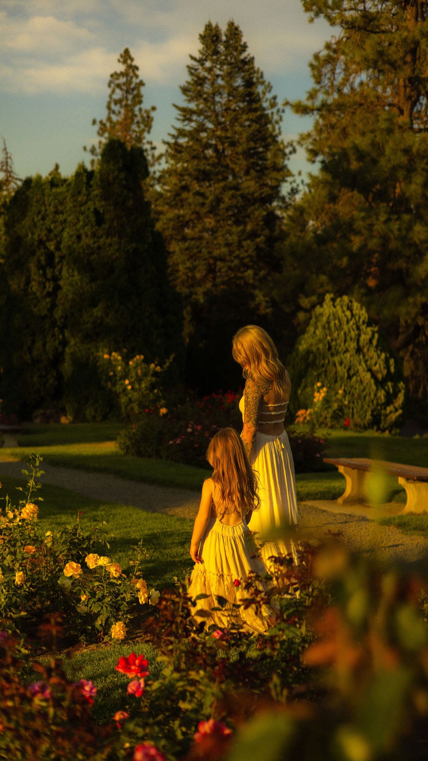 A mother and daughter in matching yellow dresses admire colorful roses in a sunlit garden surrounded by lush green trees.