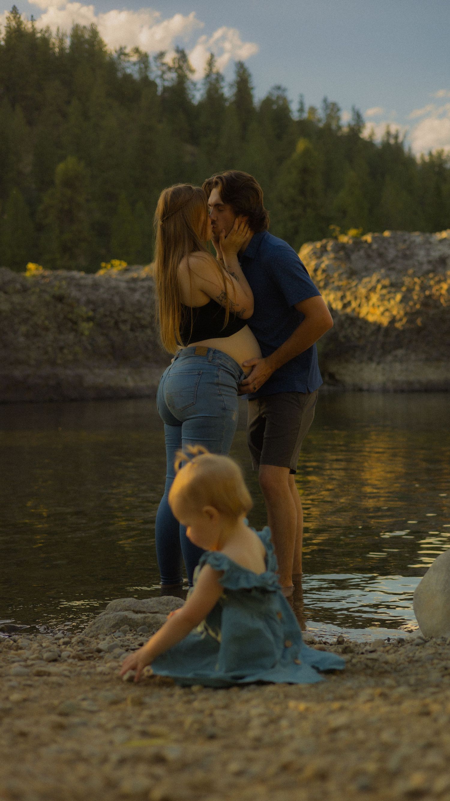 A couple shares a kiss by a lakeside while a toddler plays in the sand at golden hour.