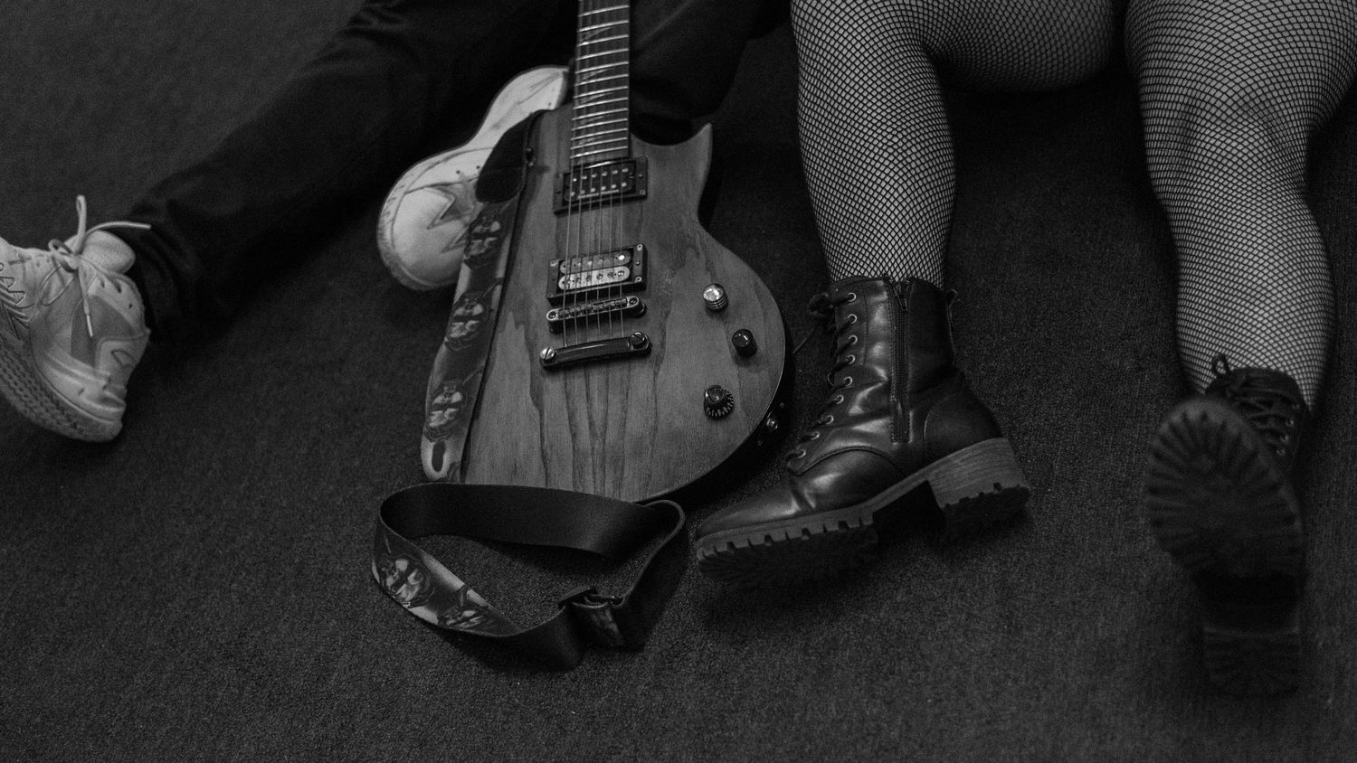 Black and white photo of two people sitting on floor with electric guitar and boots visible.