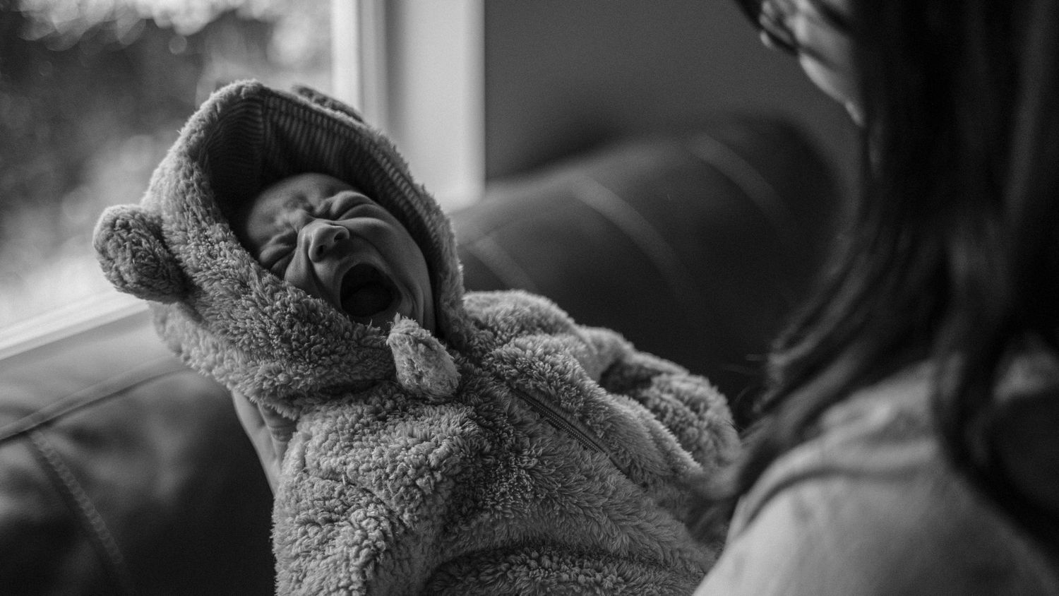 A newborn baby yawns while wrapped in a cozy gray bear-eared blanket in black and white photo.