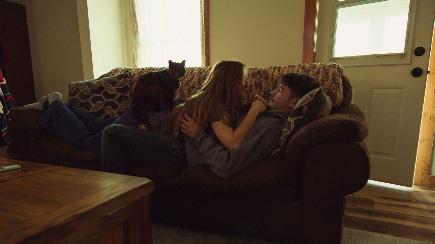 Three people relaxing together on a brown sofa with a cat perched on the back, in a cozy living room.