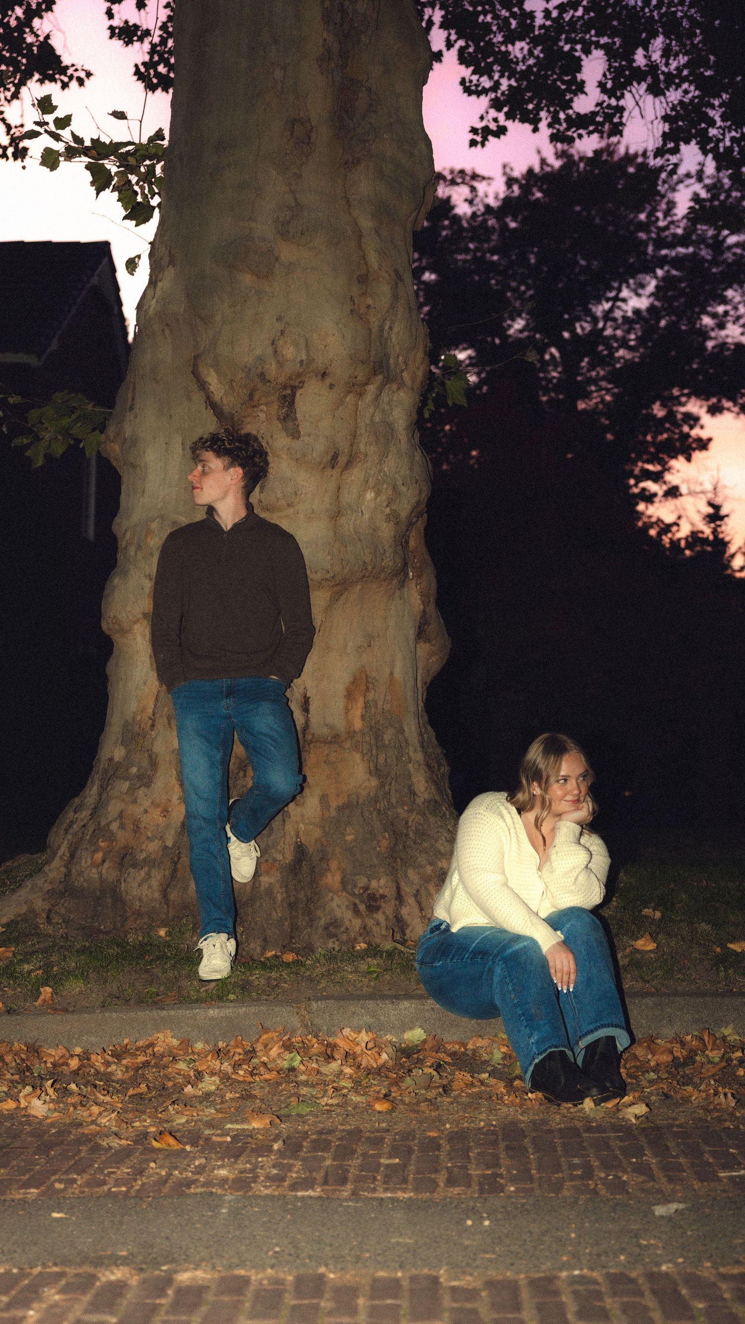 Two young people near a large tree at dusk, one standing and one sitting on a brick path surrounded by autumn leaves.