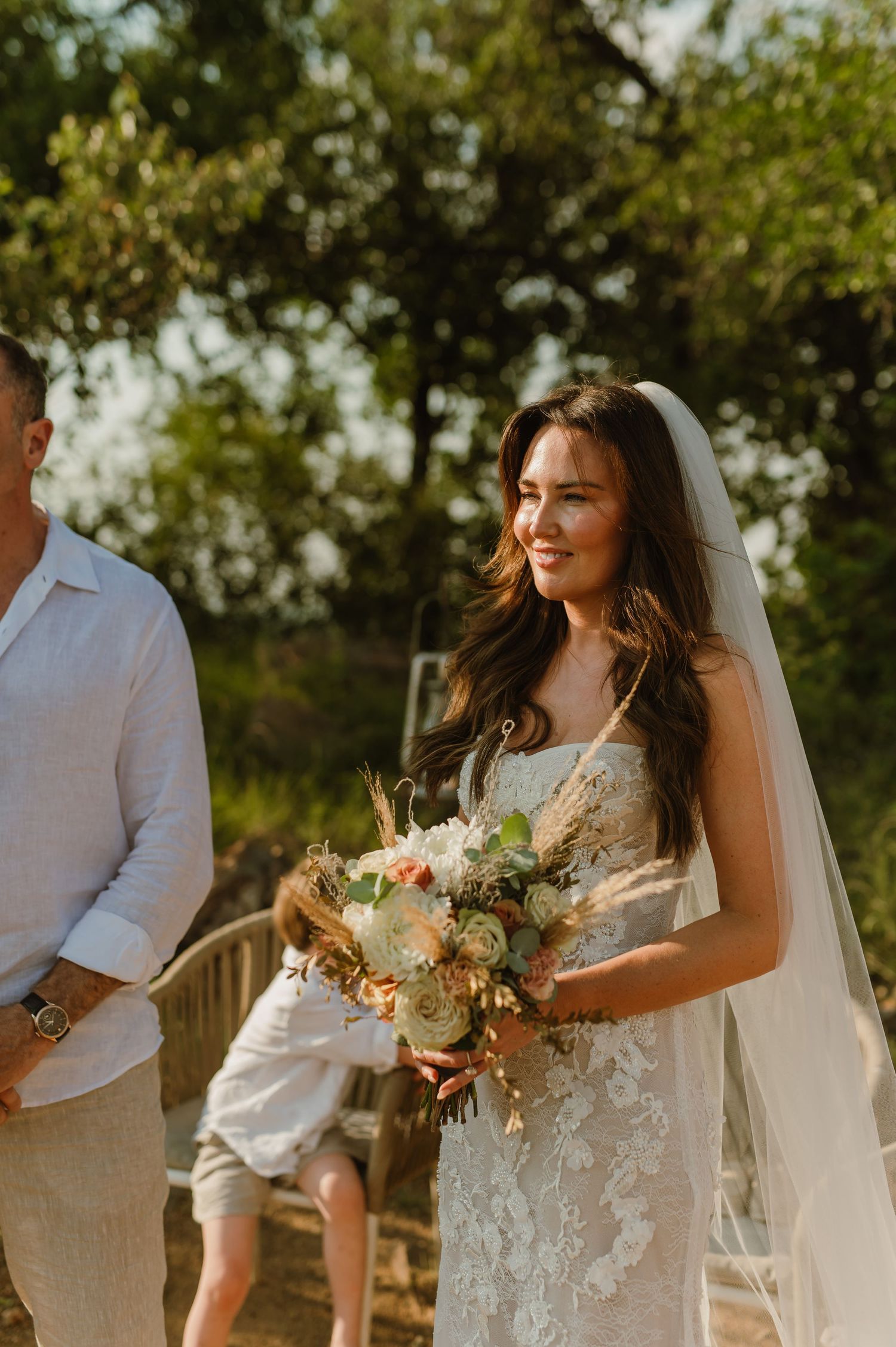 Radiant bride in lace gown and veil walking down the aisle holding a boho bridal bouquet.