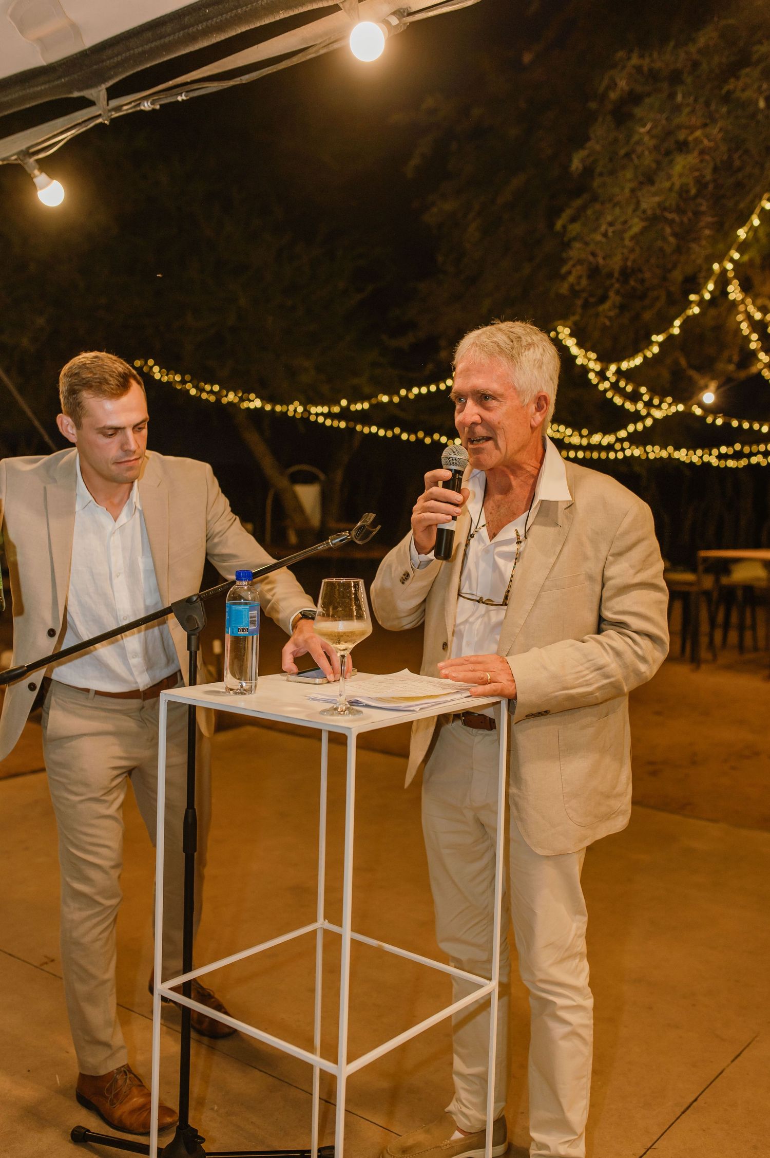 Guests in light colored suits socializing at cocktail tables during an evening wedding reception.