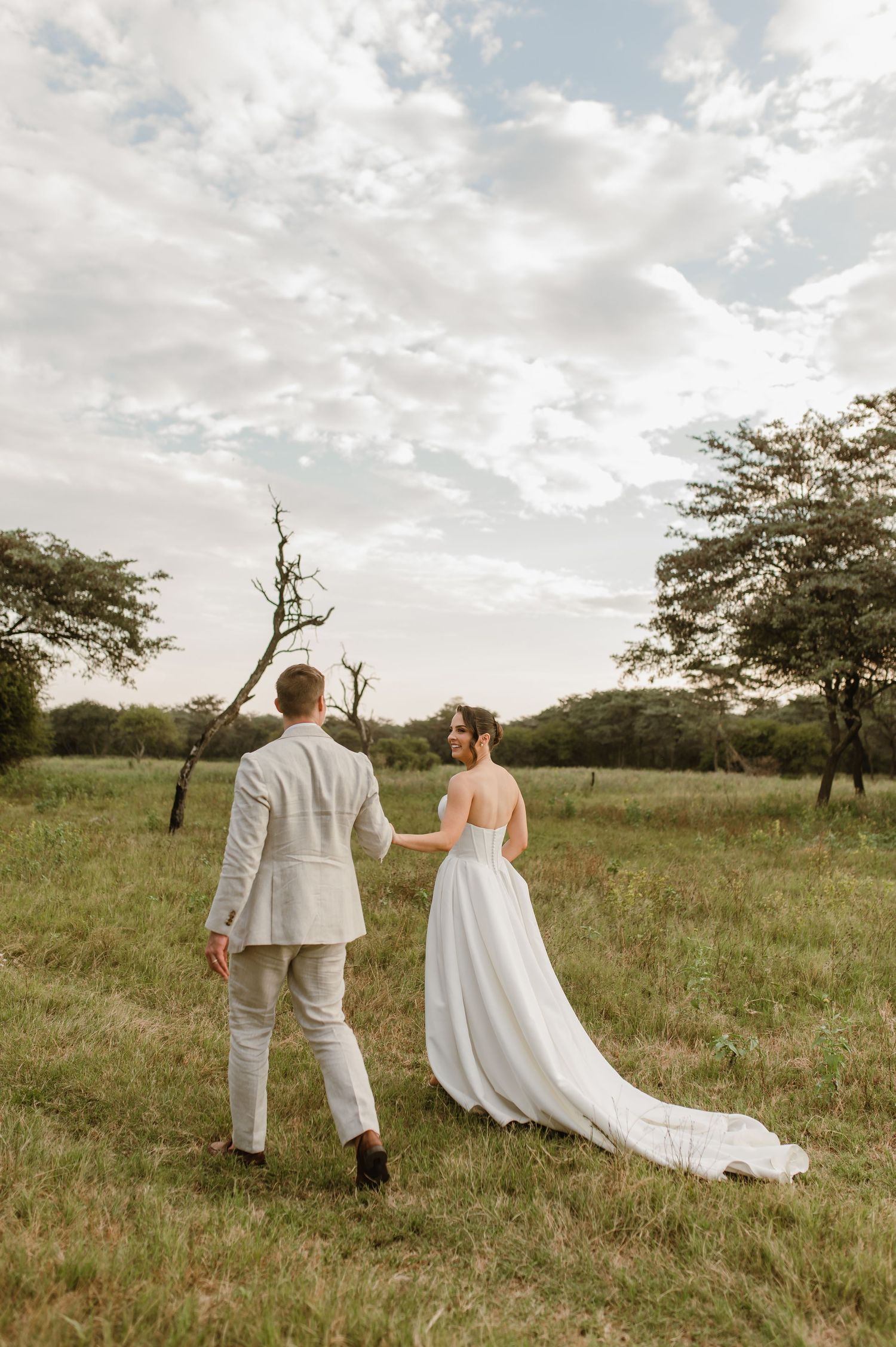 A couple in white wedding attire walks through a grassy field with scattered trees at sunset.