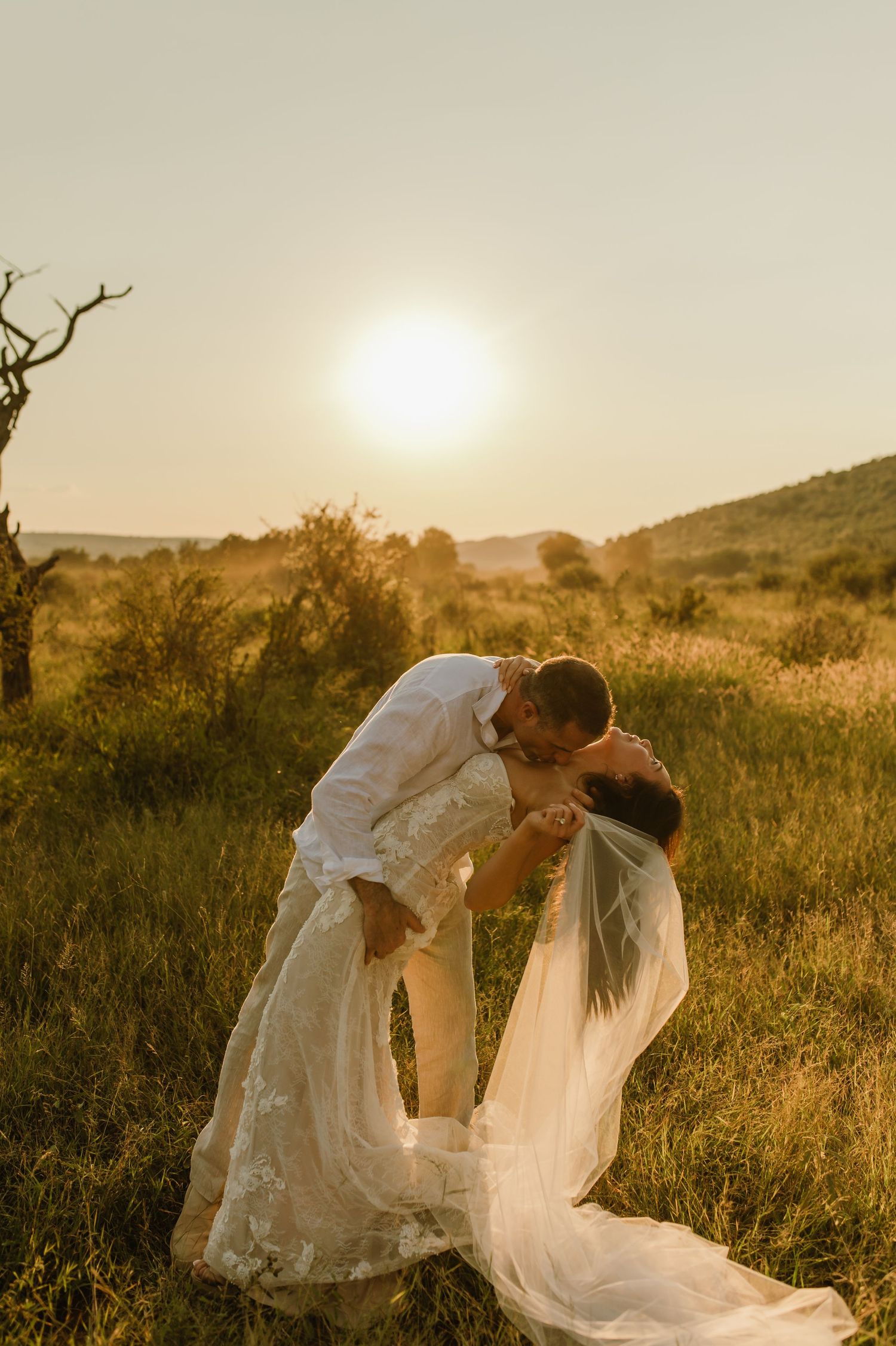 Couple shares romantic dip kiss in golden sunset meadow, bride in lace gown with veil.