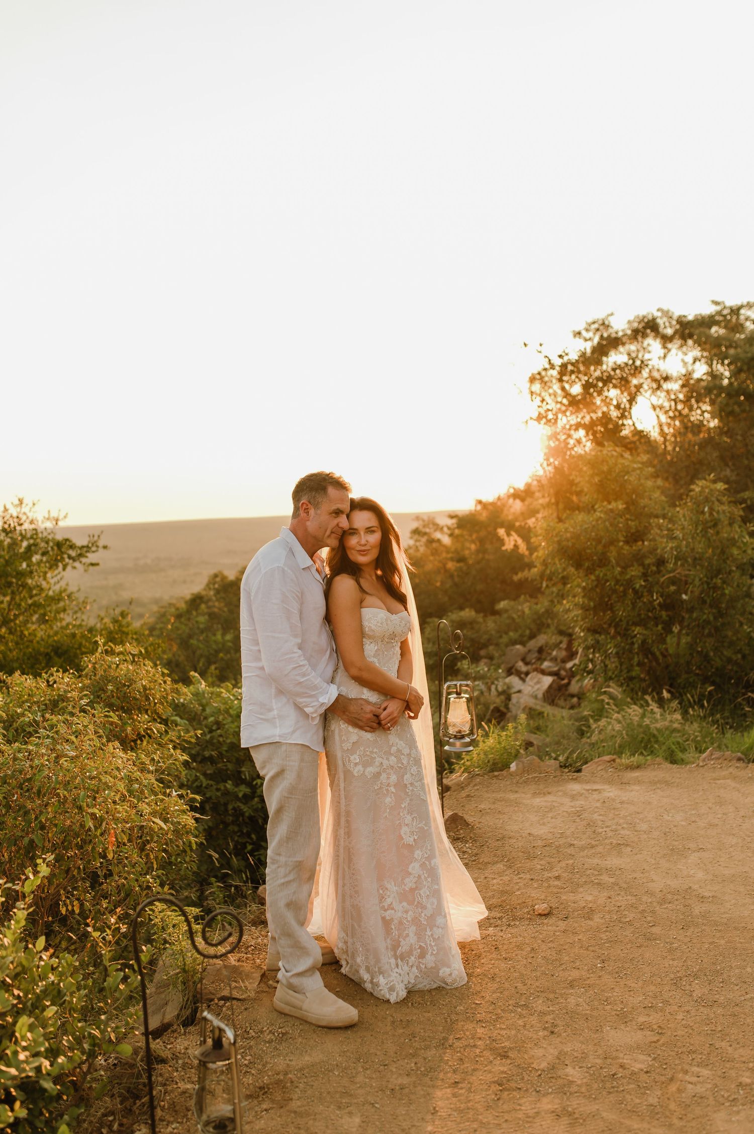 Newlyweds pose at sunset overlook, bride in lace gown with veil, groom in white linen beside lantern.