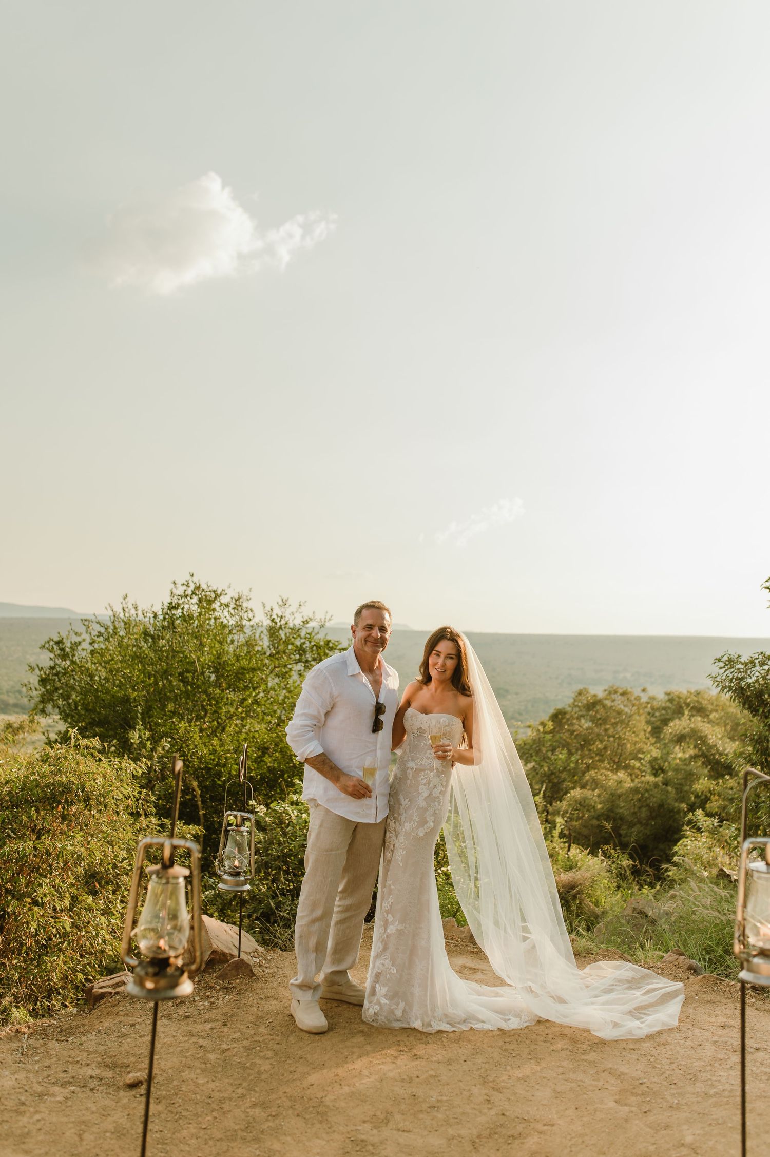 Bride and groom pose together in white attire with cathedral veil at scenic outdoor wedding ceremony.