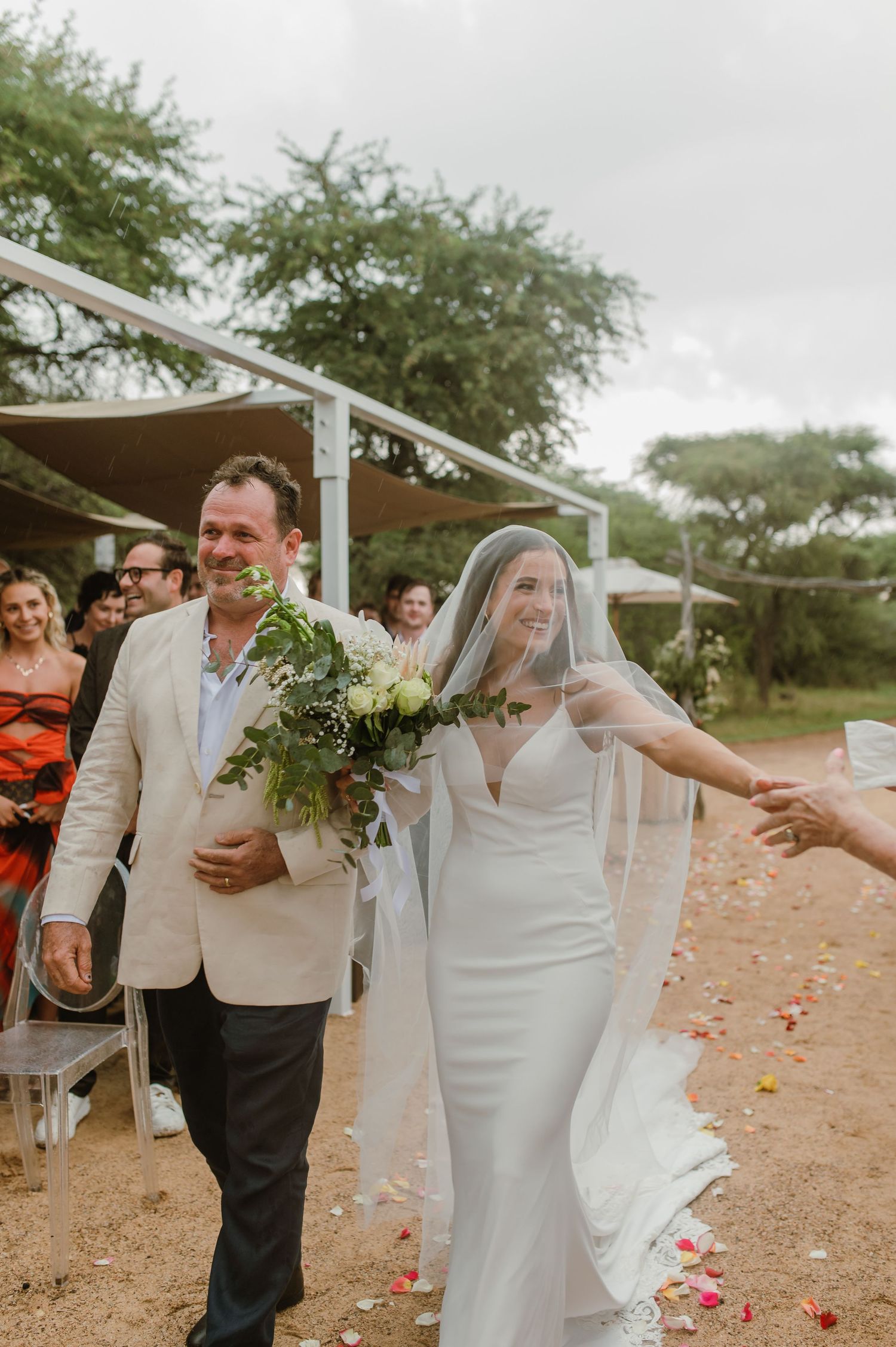 Couple walks down flower-strewn path under white canopy during outdoor ceremony.