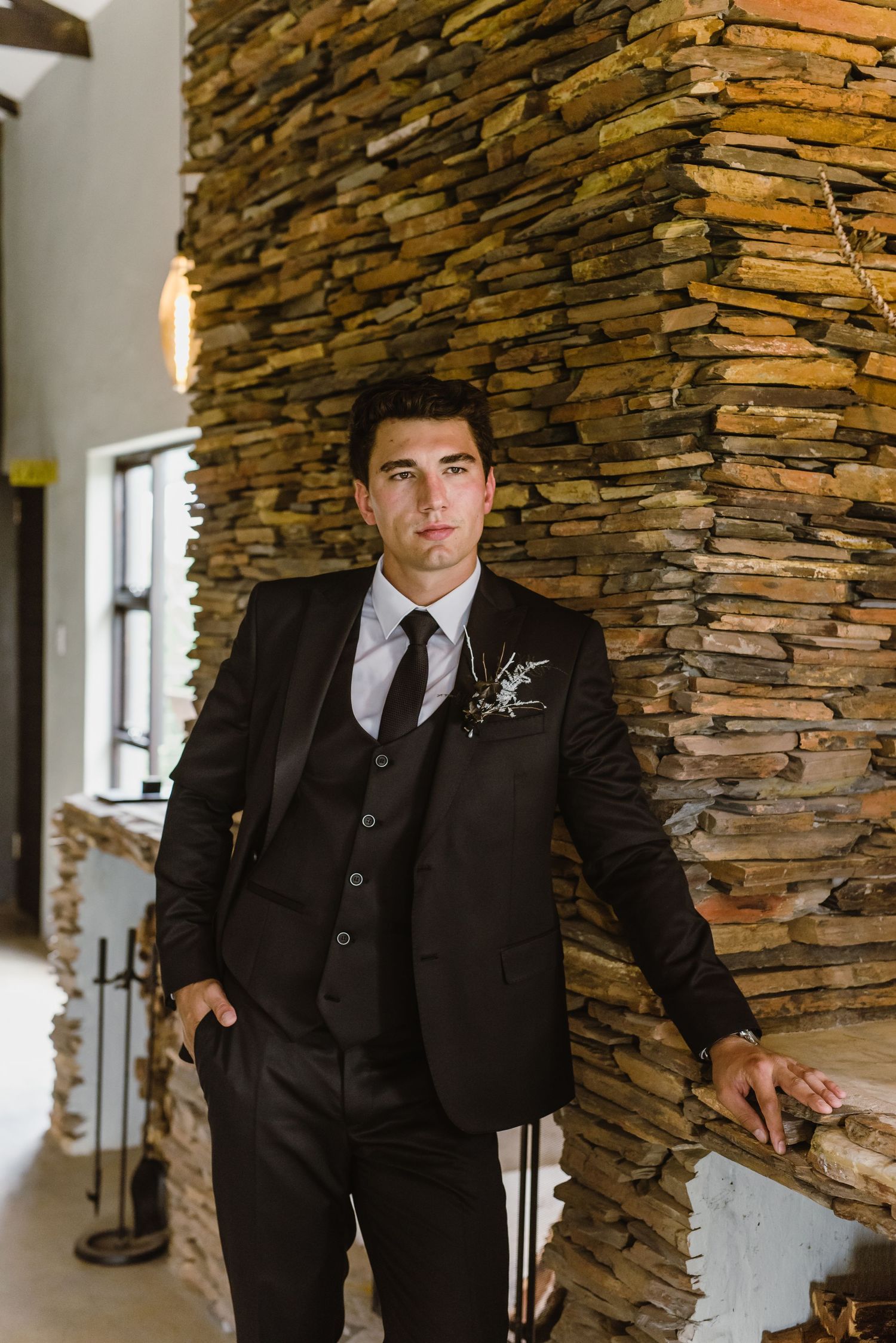 Black suit-clad figure poses dramatically against rustic stacked stone wall in modern venue.