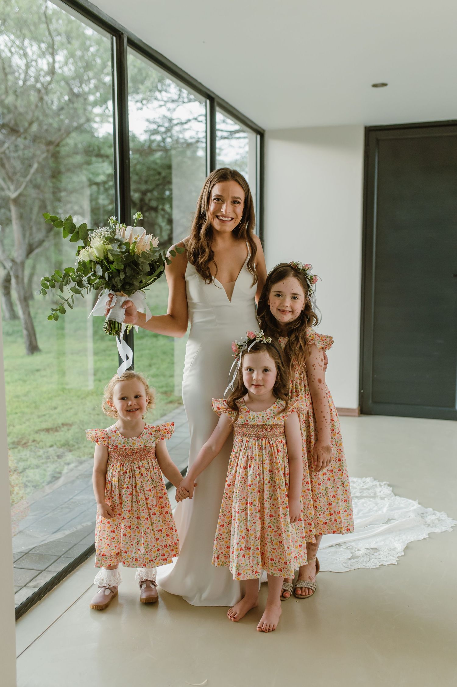 Two small children in floral dresses join someone in a white wedding gown for a playful moment.