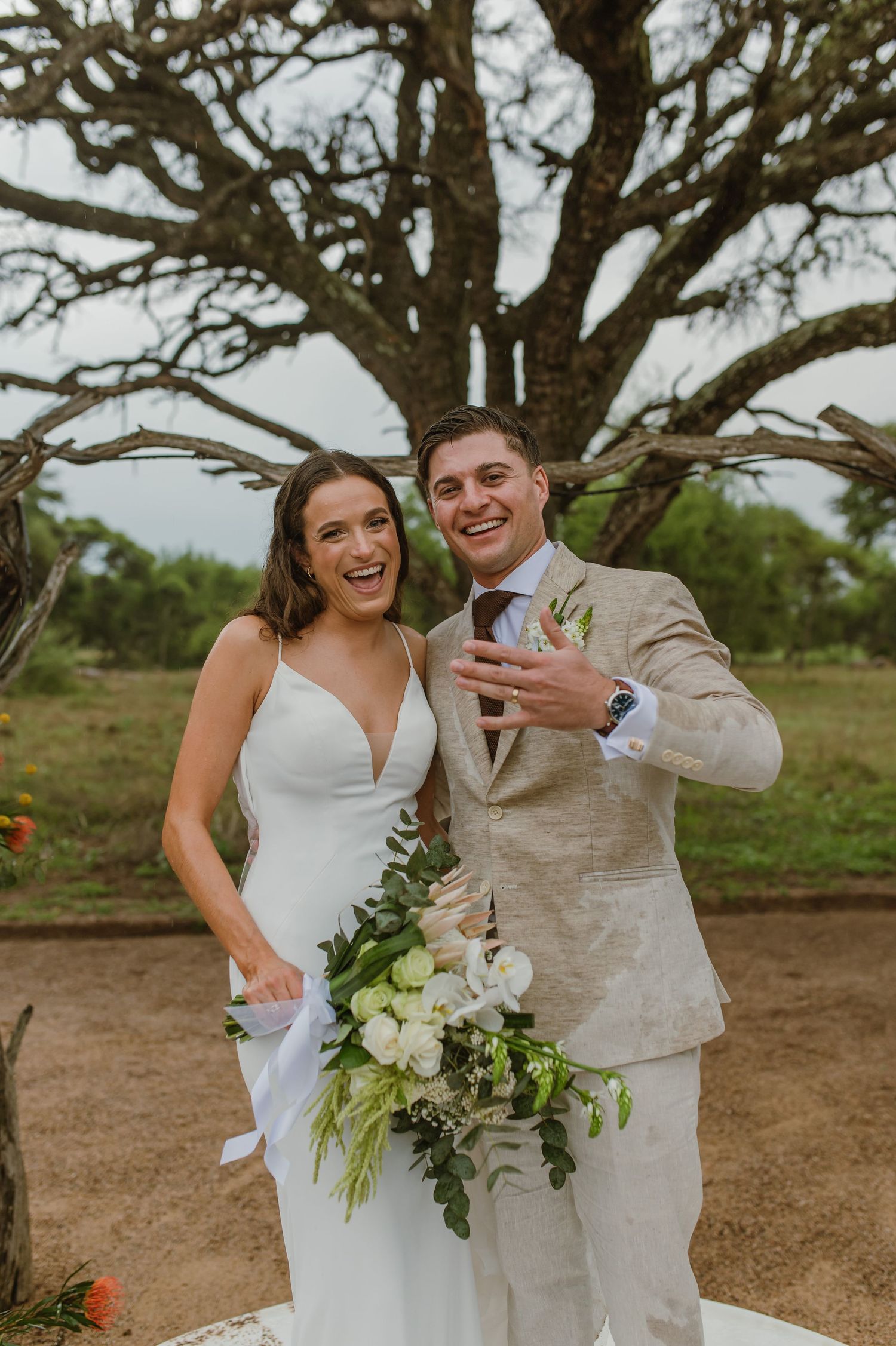 Joyful wedding couple celebrating under a large tree in an outdoor setting.