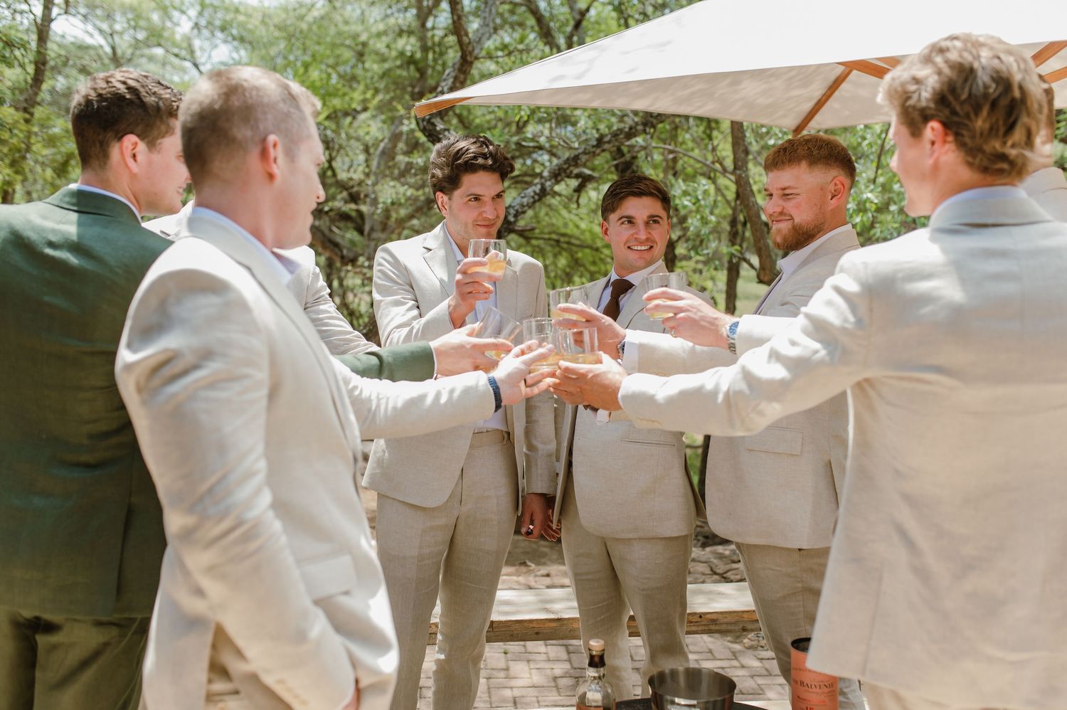 Groomsmen in matching light suits raise their glasses in a celebratory toast outdoors.