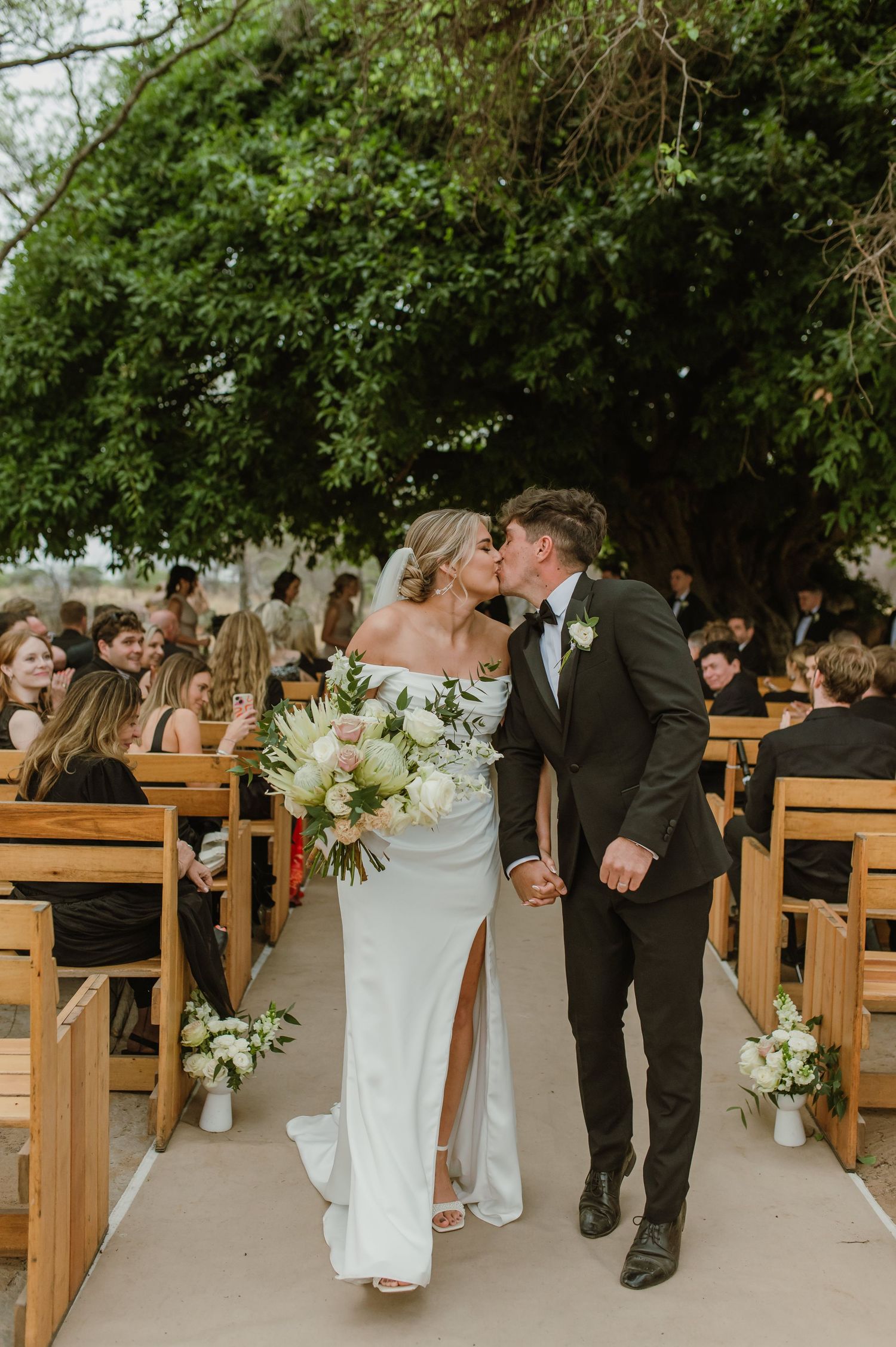 Wedding party walks down aisle between wooden pews under a natural tree canopy.