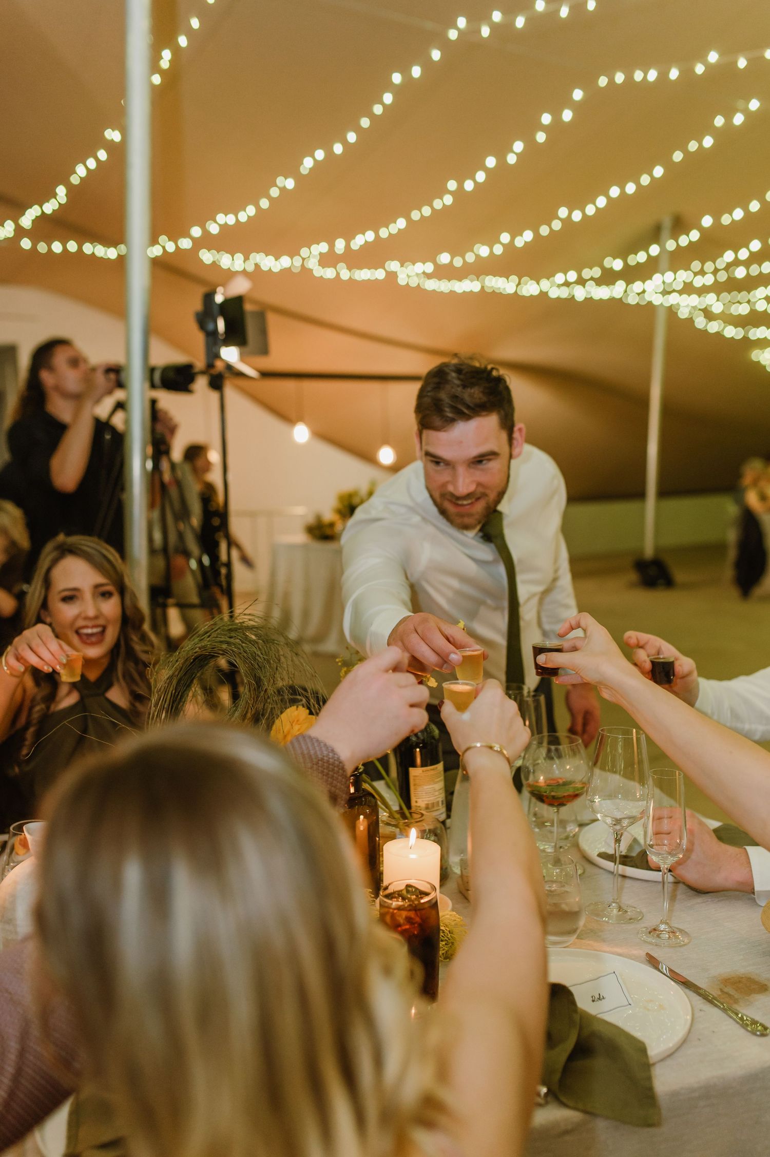 Wedding guests raising glasses in a toast under twinkling string lights at reception.